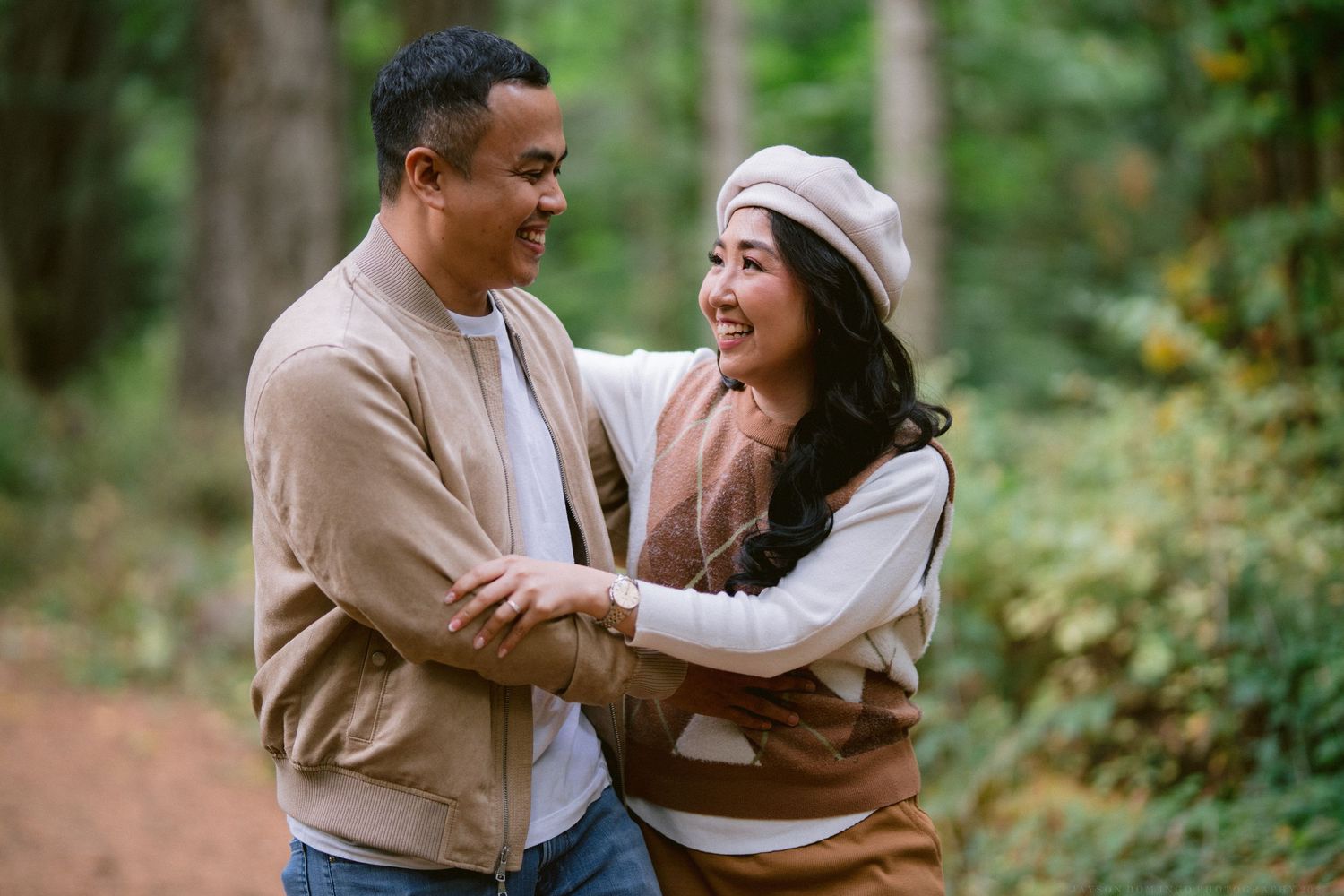 A couple in casual attire embraces while sharing joyful laughter in a scenic outdoor forest setting.