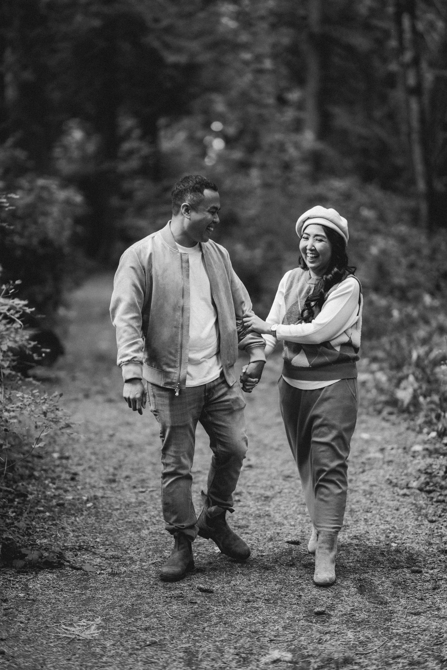 A couple walks together and laughs on a forest trail in casual fall attire.
