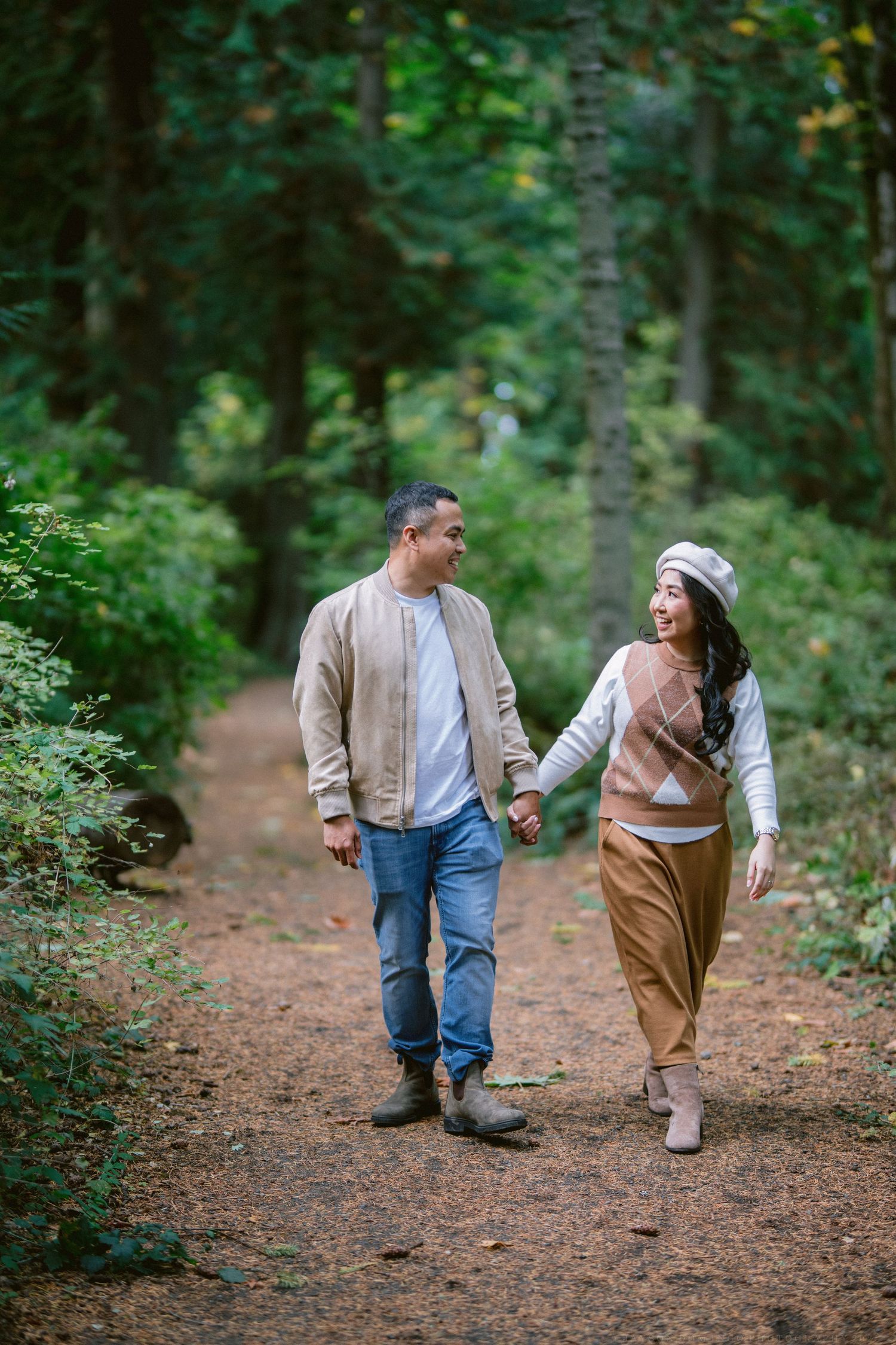 Couple walking hand in hand along a forest trail covered in fallen leaves.