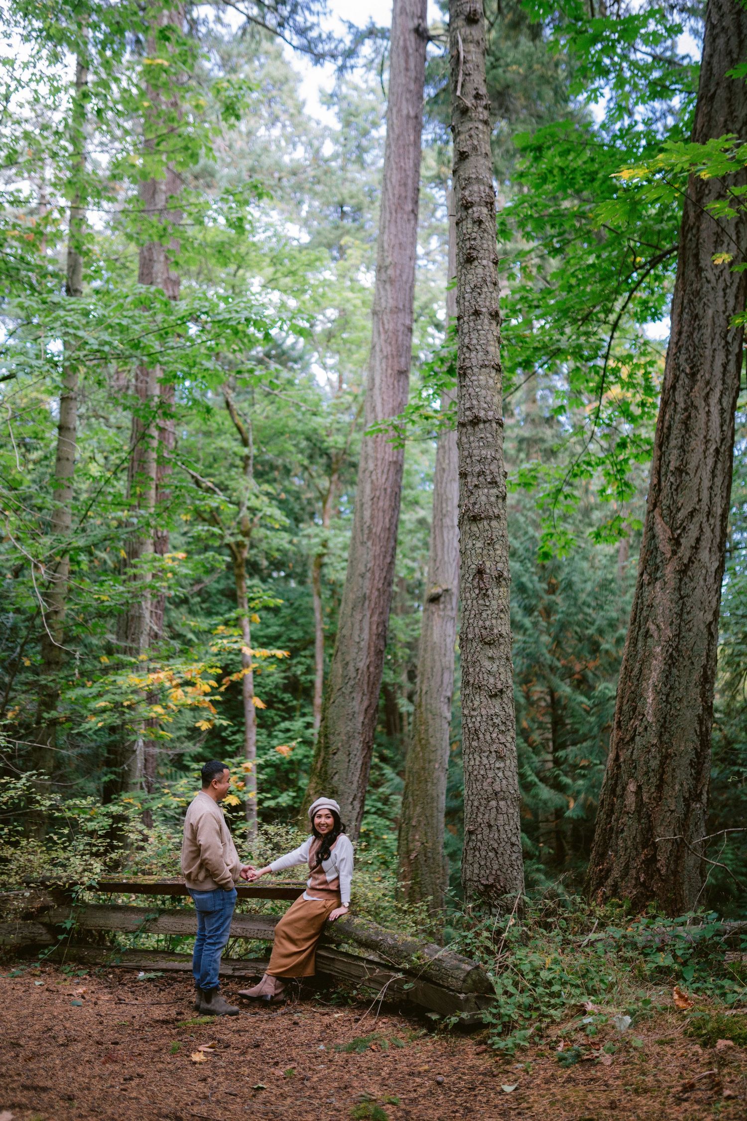 Two people walking together through a lush evergreen forest with tall trees and green undergrowth.