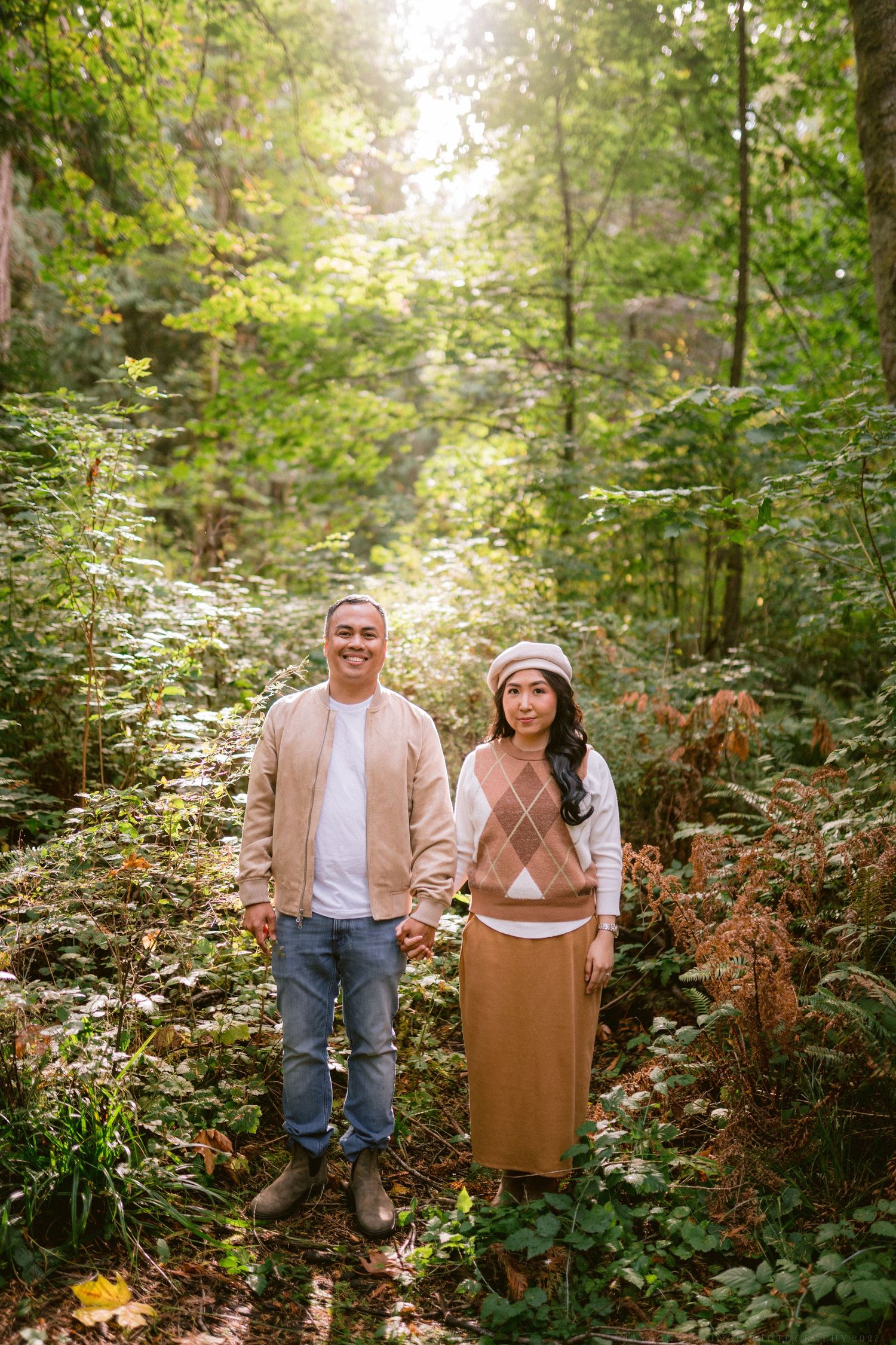 A couple poses together in a lush forest setting wearing coordinated beige and brown autumn outfits.