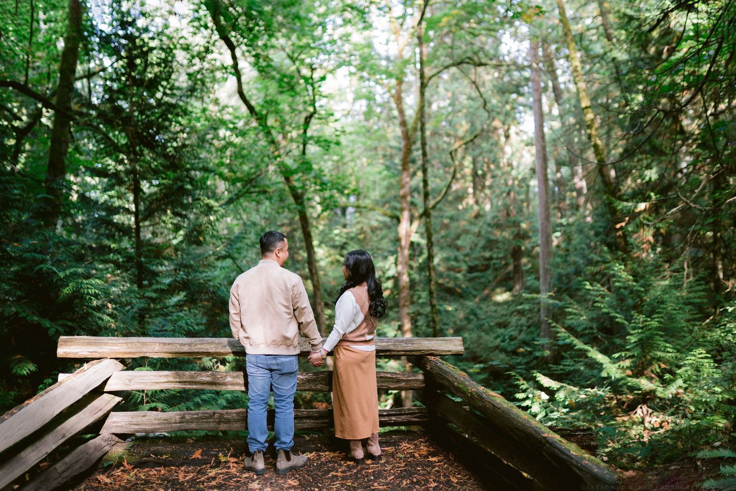 Two people walk along a wooden boardwalk path winding through a lush green forest on a summer day.