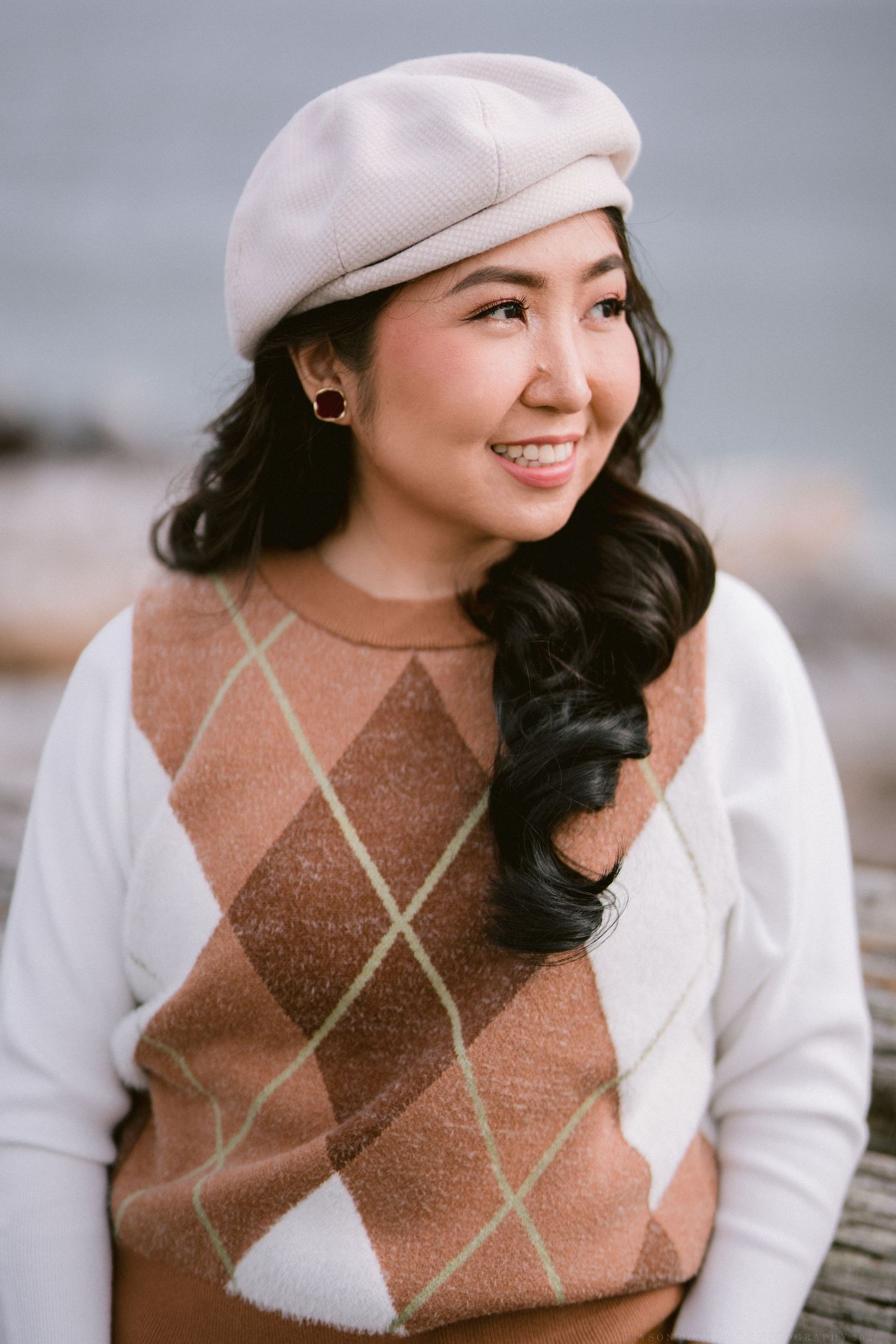 A person wearing a brown argyle sweater vest and white cardigan poses outdoors with a warm smile.
