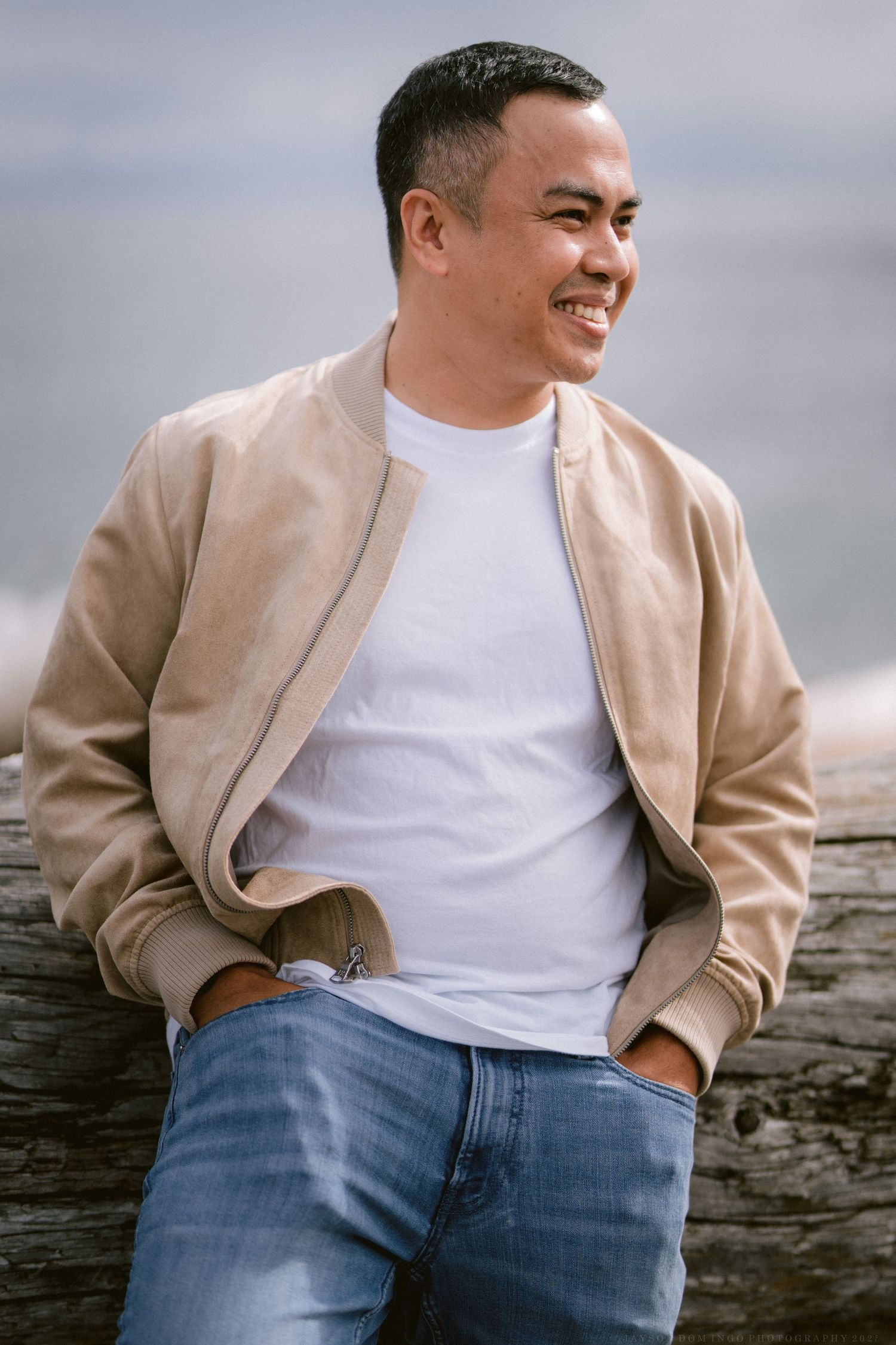 A person wearing a beige bomber jacket and white t-shirt poses casually against a coastal backdrop.