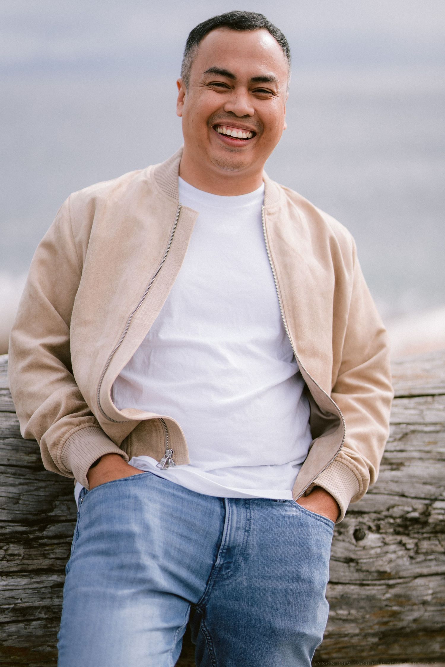 Person in beige jacket and jeans sitting casually on weathered wooden fence.