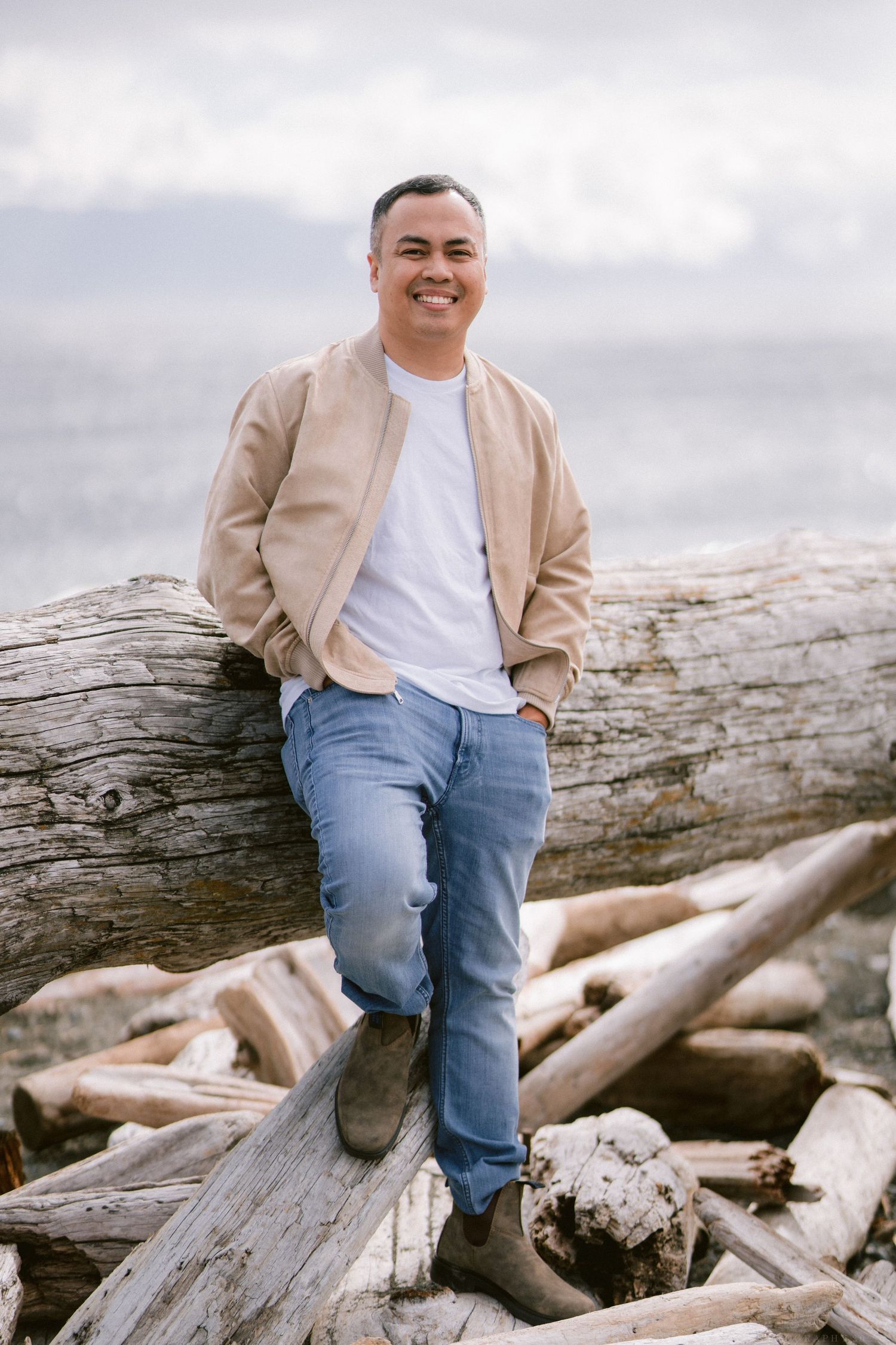 Person in casual attire sits on driftwood logs at a beach during an overcast day.