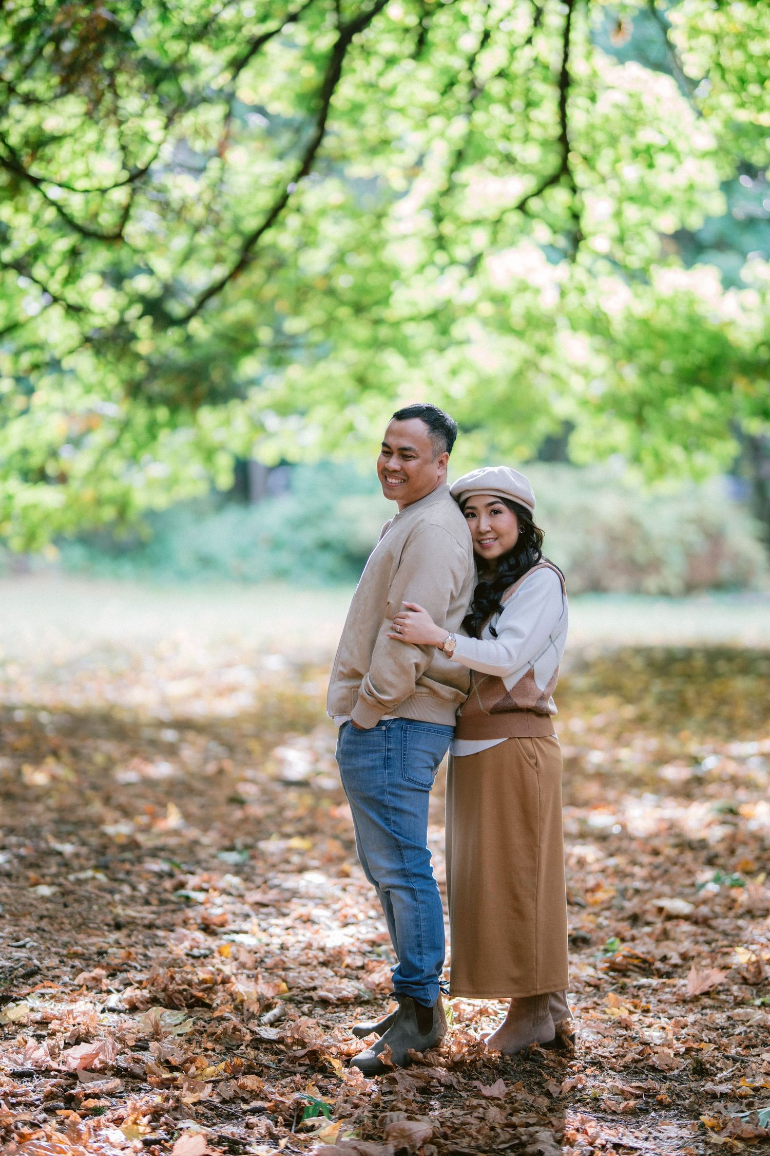 A couple shares a tender embrace while standing on fallen autumn leaves under green trees in a park.
