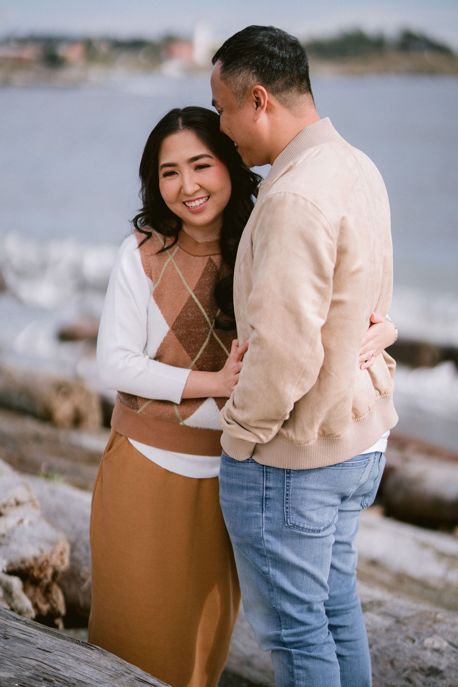 A couple shares an intimate moment by the ocean, wearing coordinated beige and brown outfits.