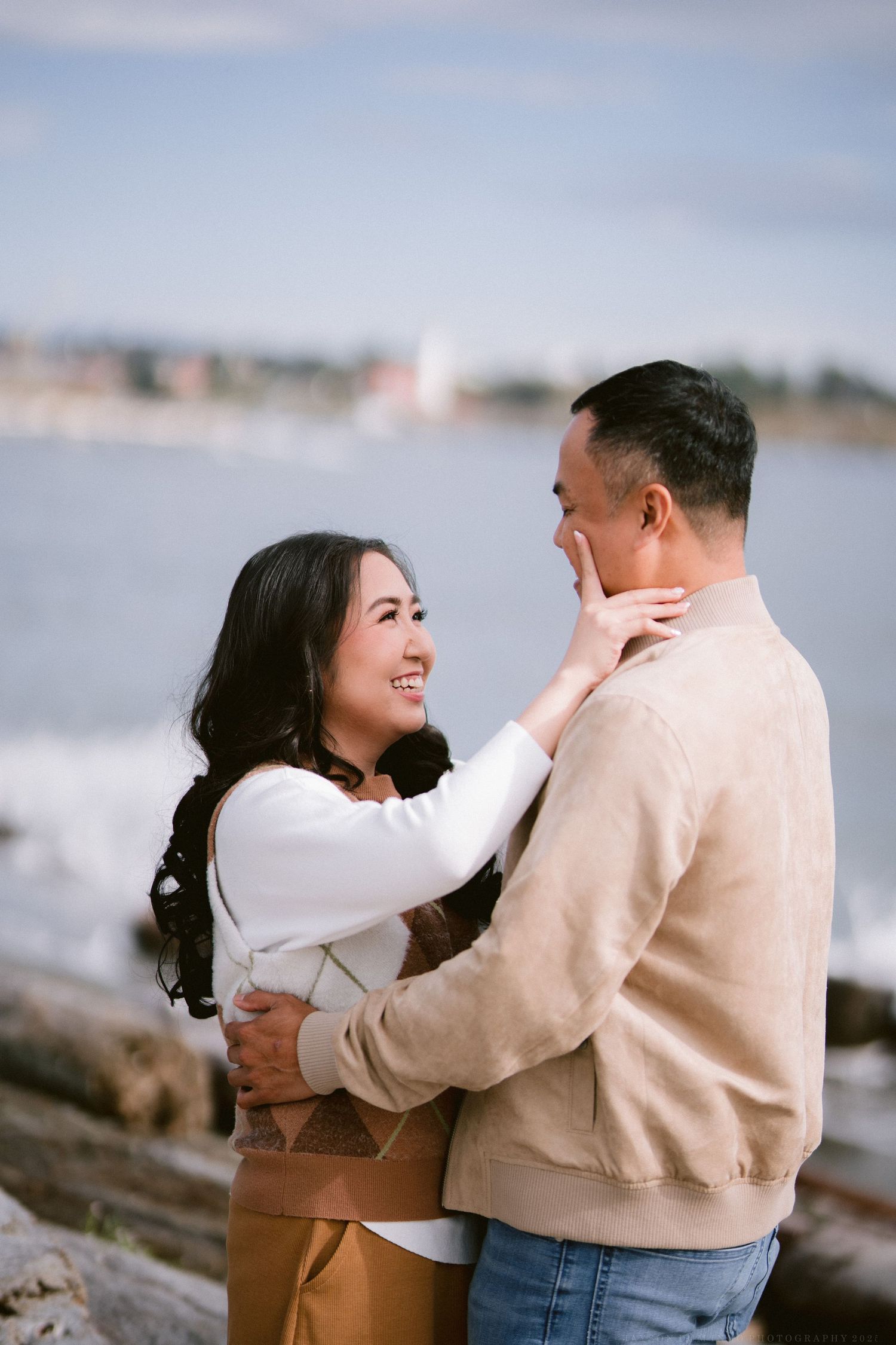 A romantic moment captured between a couple in light colored sweaters on a coastal shoreline.