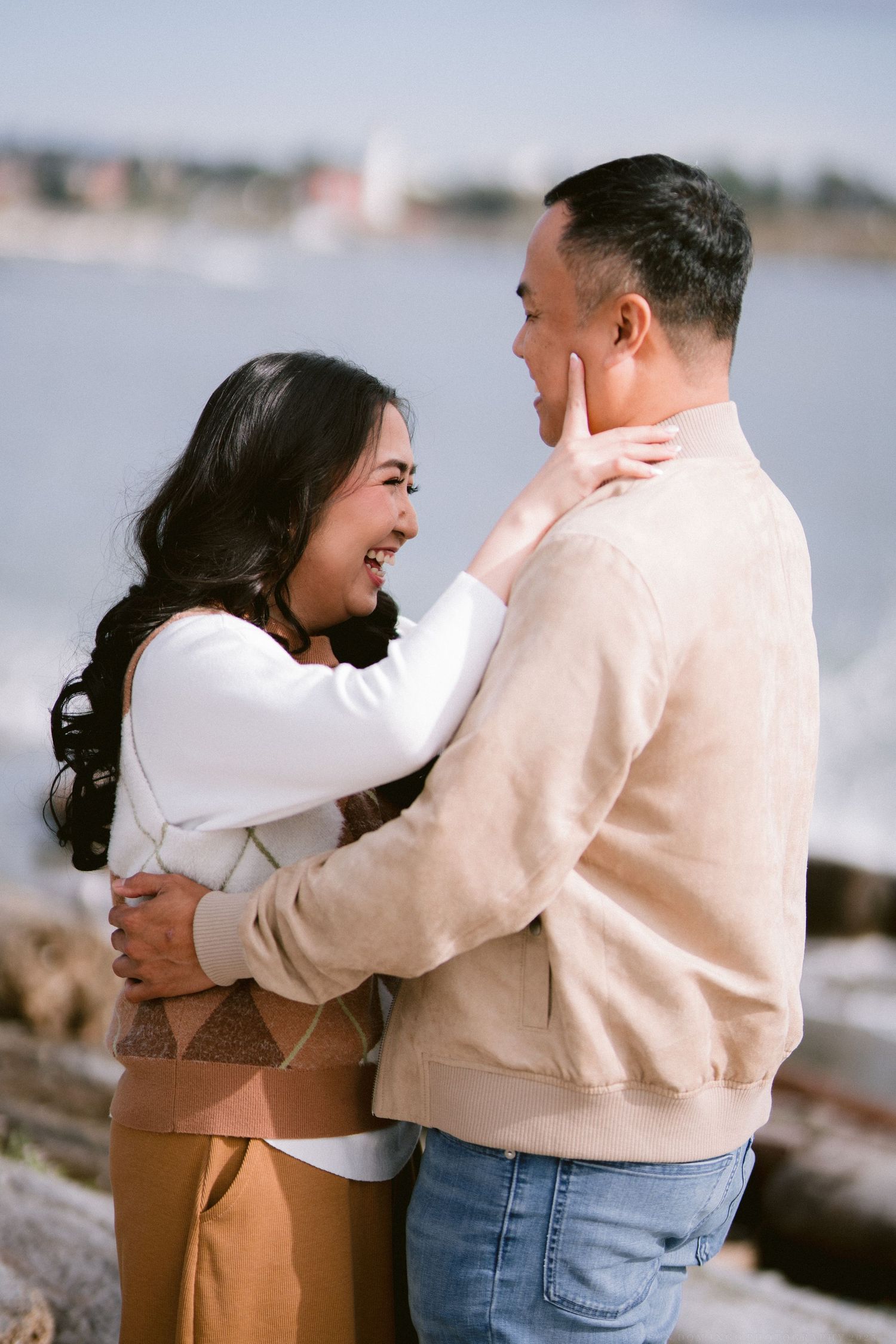 A couple sharing a joyful embrace while wearing matching light-colored sweaters by the waterfront.