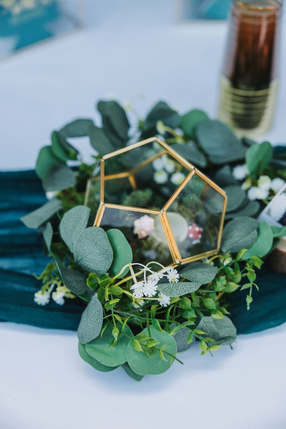 Geometric gold ring holders displayed on eucalyptus leaves and delicate white flowers create an elegant wedding detail setup.
