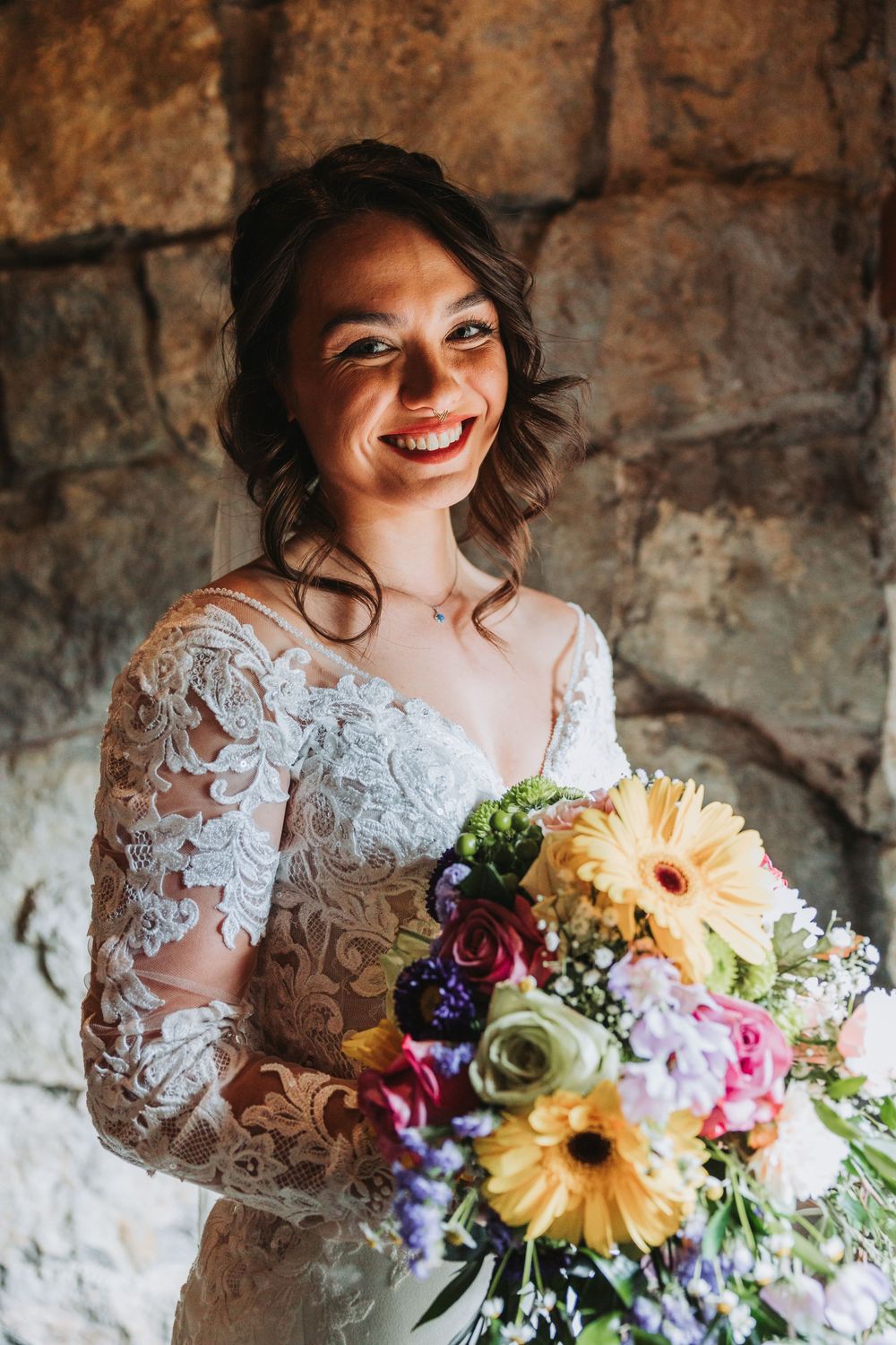 A joyful bride in lace sleeves holds a vibrant bouquet with sunflowers, roses and purple blooms against a rustic stone wall.