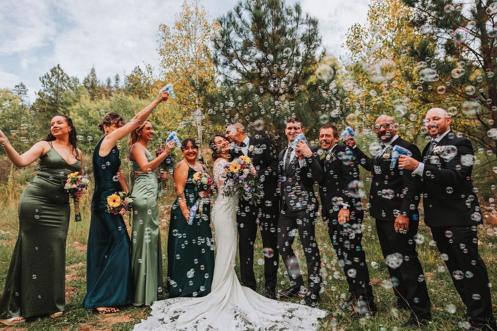 Wedding party celebrates outdoors with confetti in a forest setting while wearing formal attire.