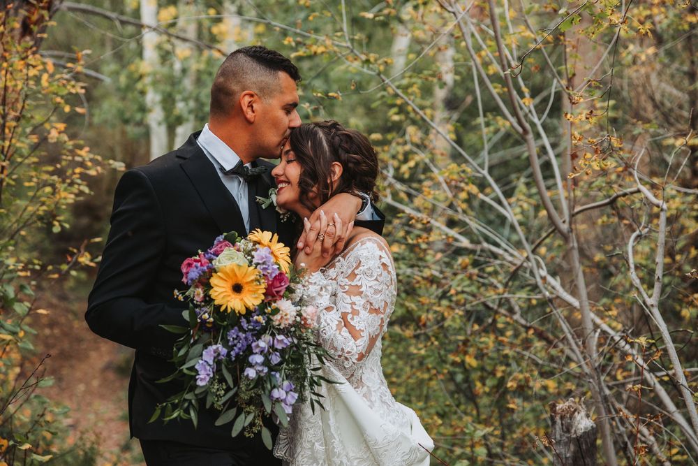 Romantic couple shares a tender kiss in a wooded outdoor setting surrounded by autumn foliage and holding floral bouquets.