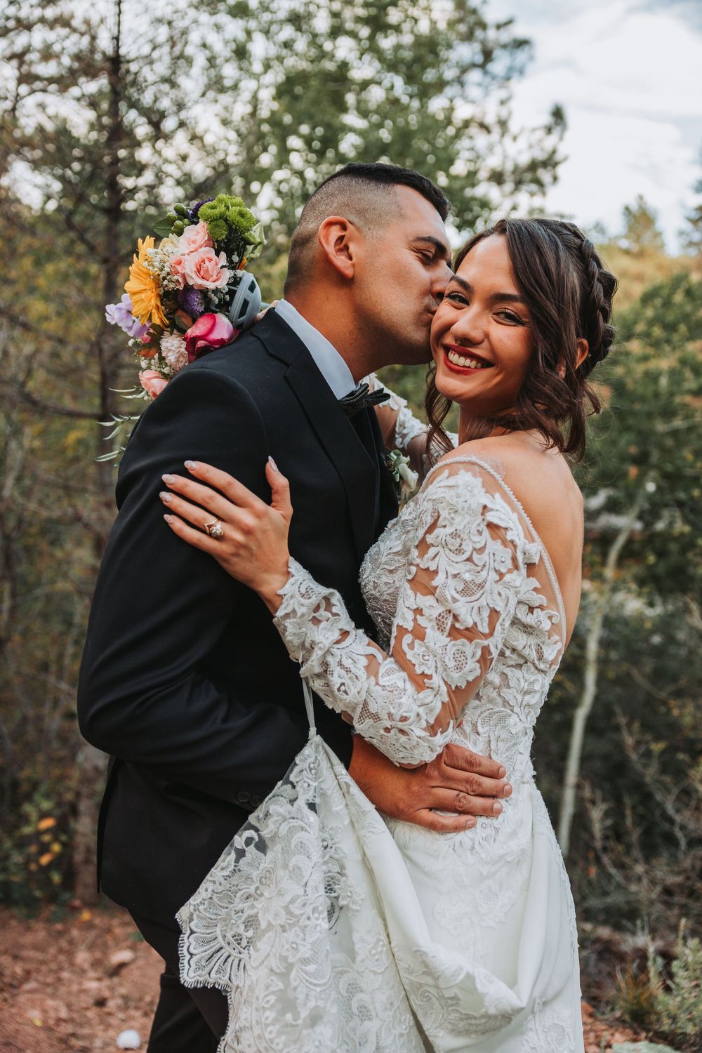 A romantic outdoor wedding photo showing a couple embracing in lace dress and black suit among autumn trees.