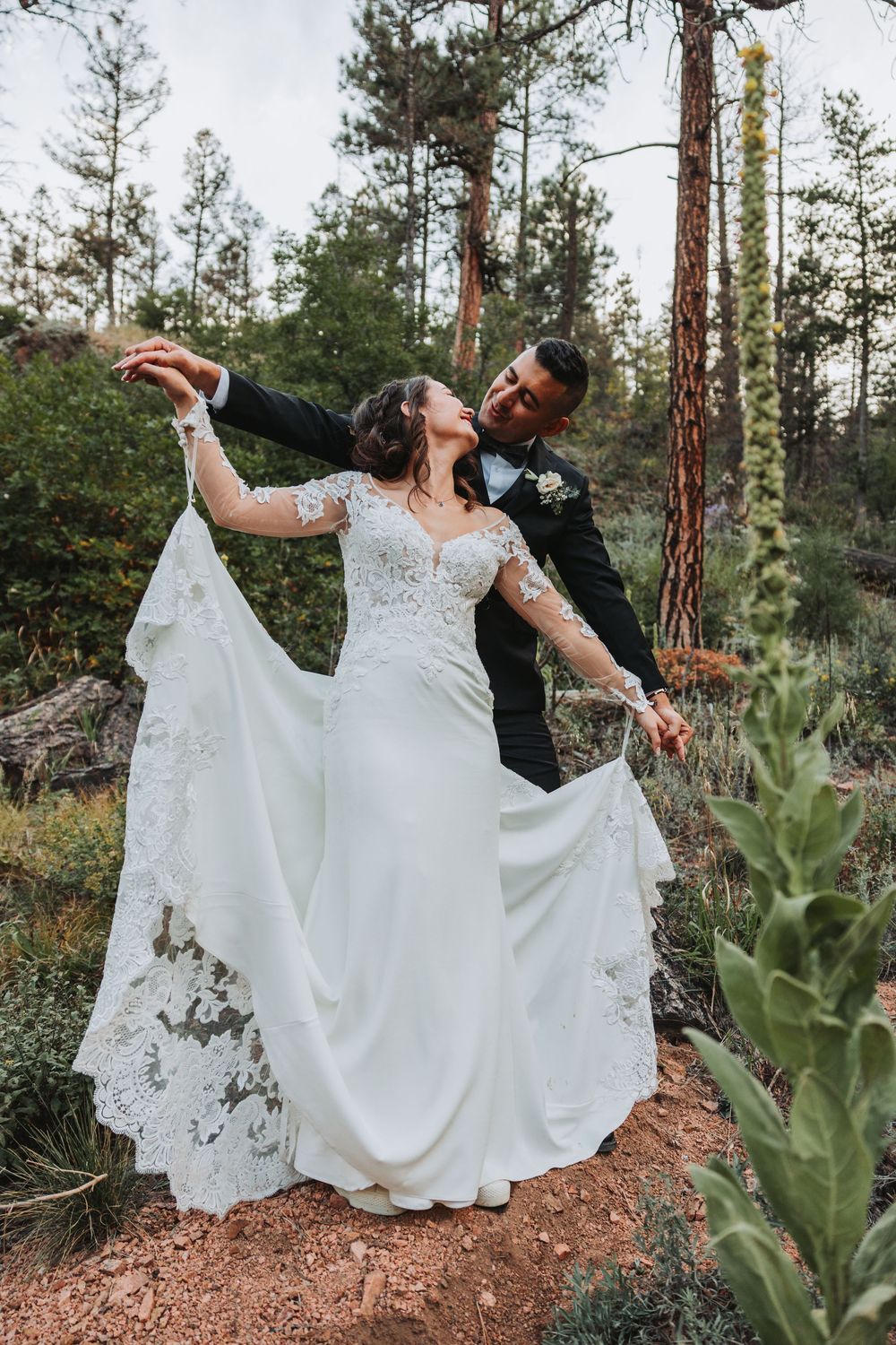 A couple in wedding attire dances together on a mountain trail surrounded by tall pine trees at sunset.