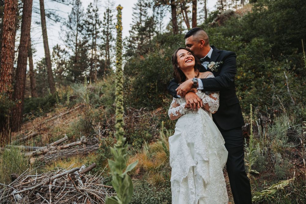 A couple in wedding attire embrace on a wooded hillside with tall pine trees and wildflowers in the background.