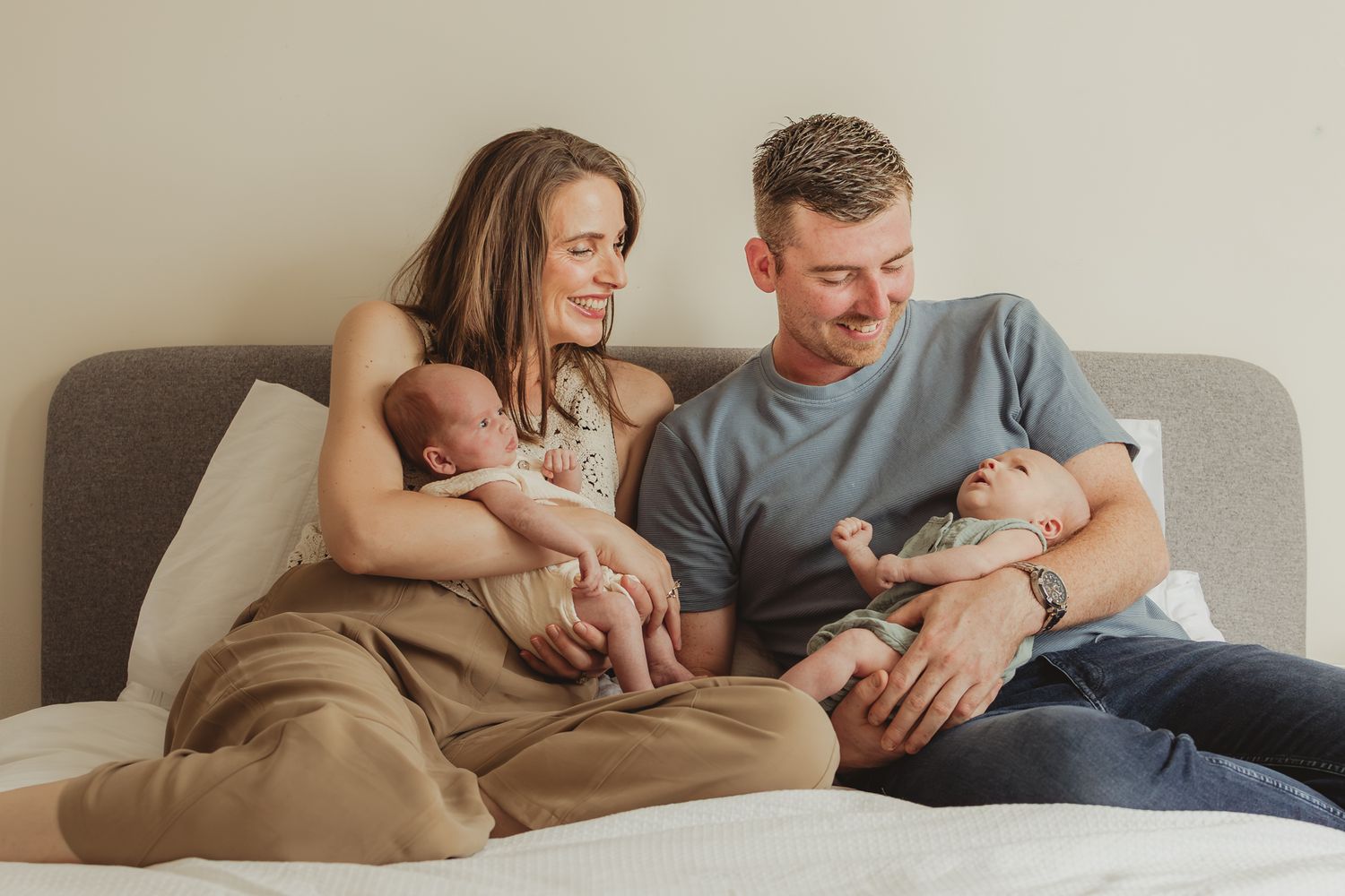 Parents sitting on gray couch holding twin newborn babies in their arms while sharing a tender family moment.