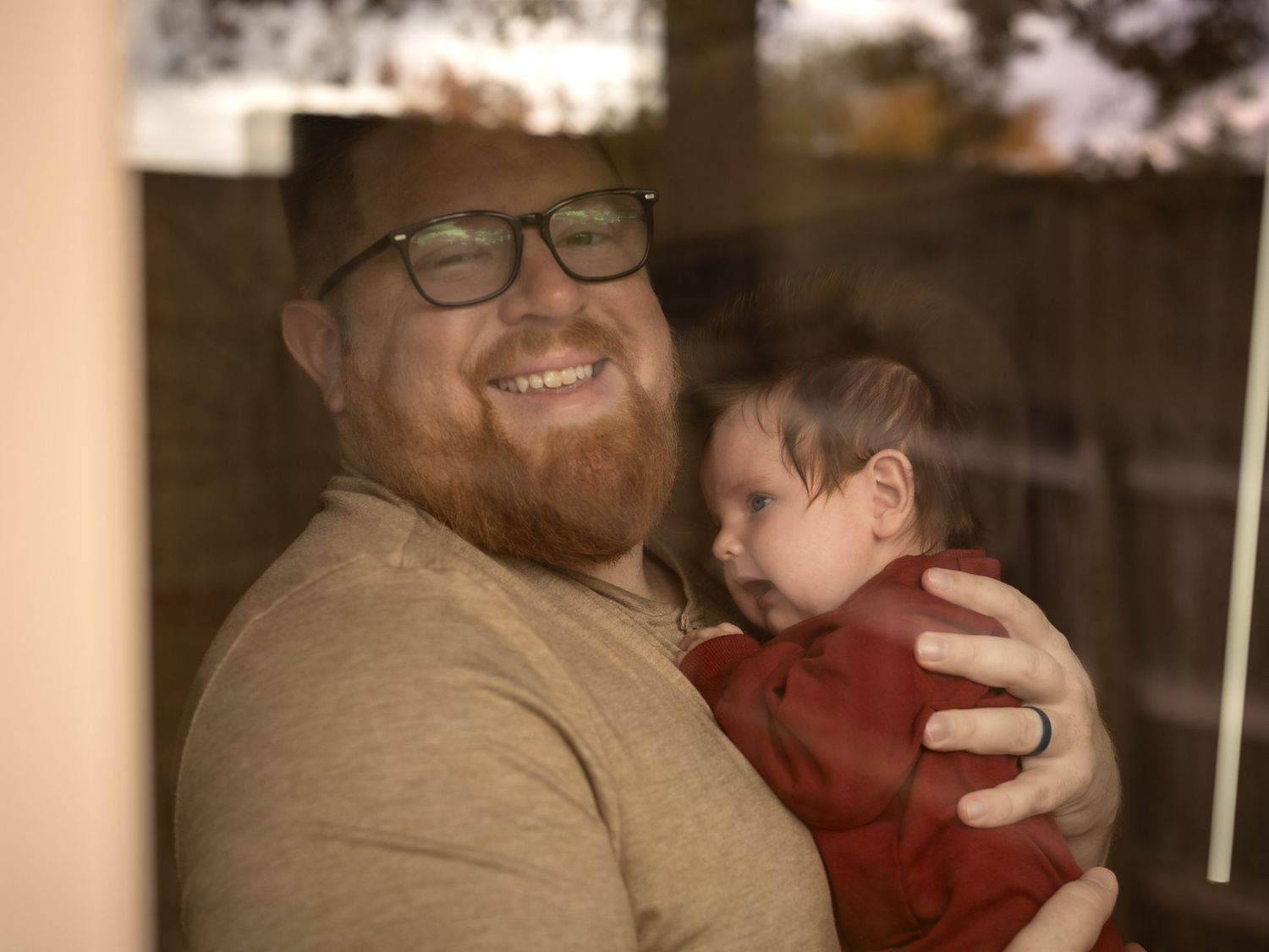 A bearded person in glasses holds a small baby wearing red clothing in a warm, intimate indoor setting.