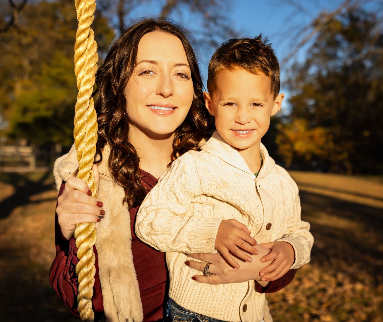 A parent and child smile together while holding a rope swing in a sunlit autumn park.
