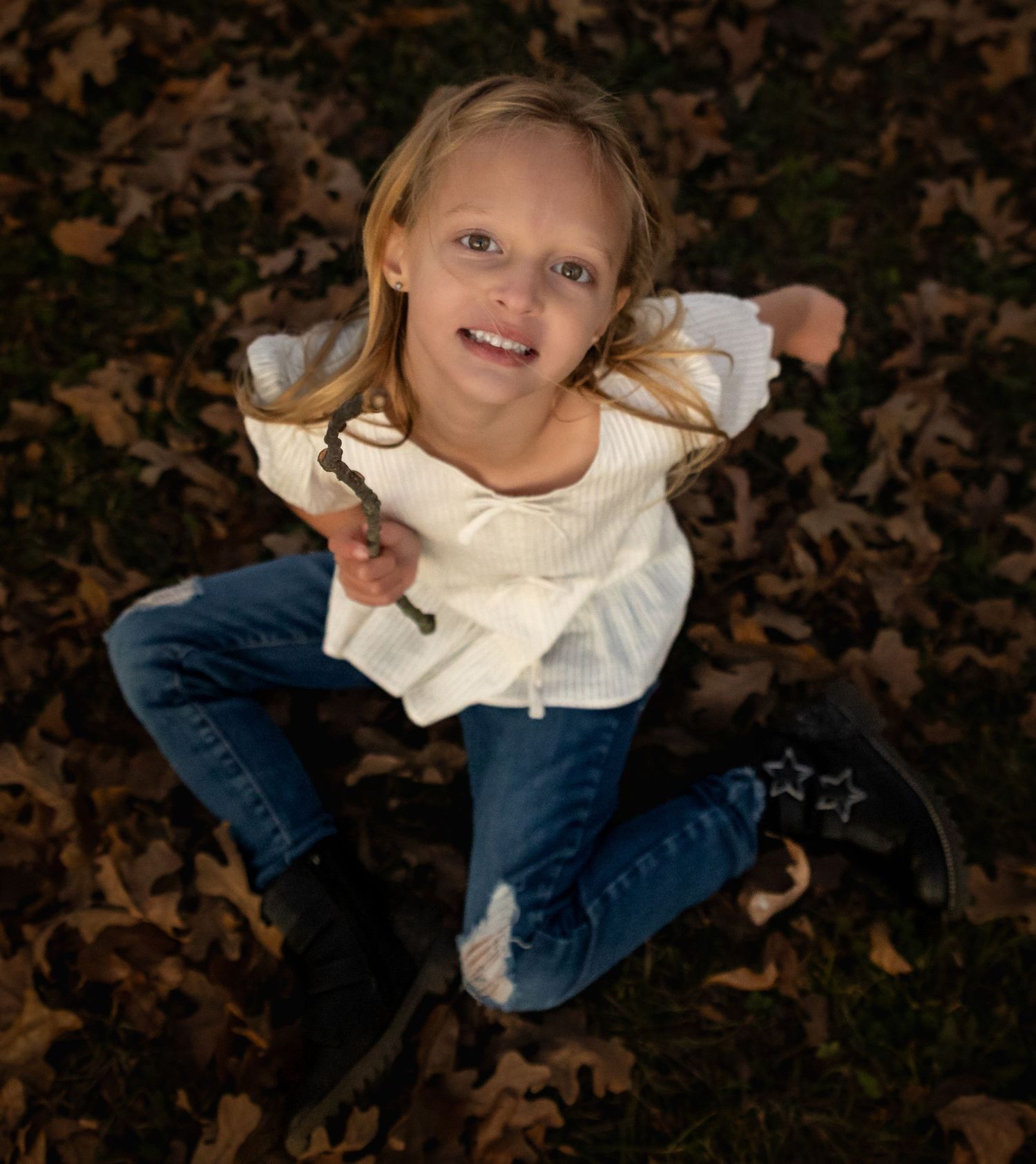 Child in blue jeans and white sweater lies on fallen autumn leaves while smiling at the camera.