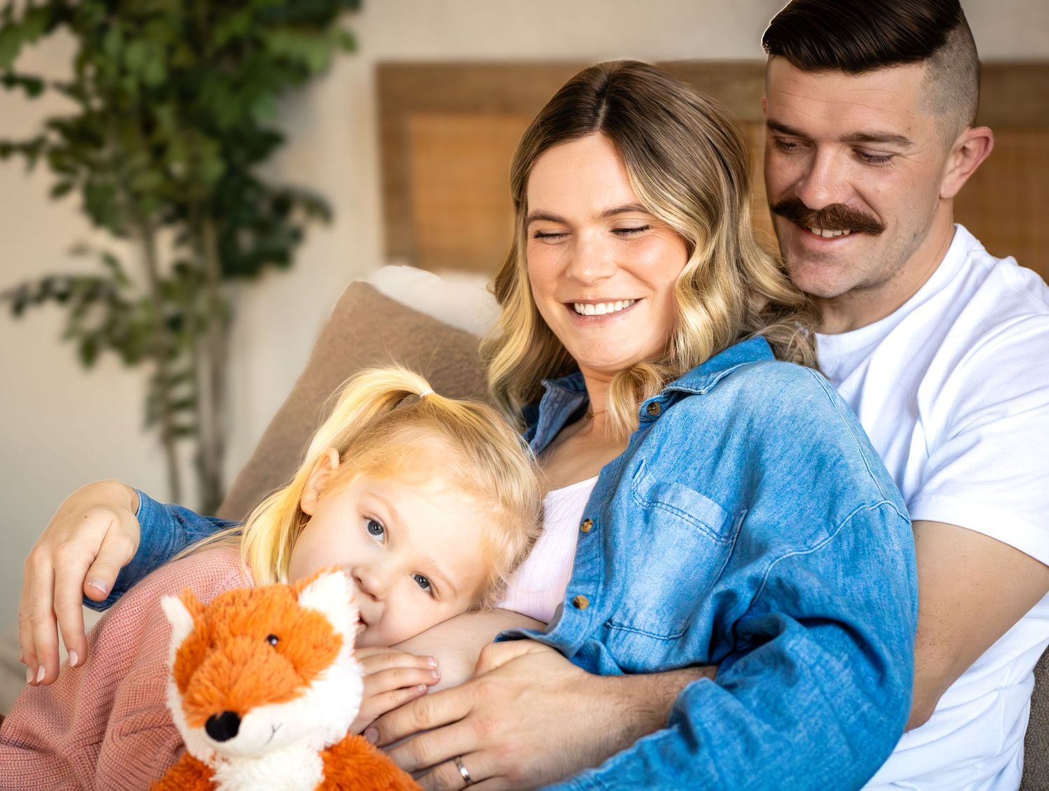 A family relaxes together on a couch at home with a stuffed fox toy and blue blanket.