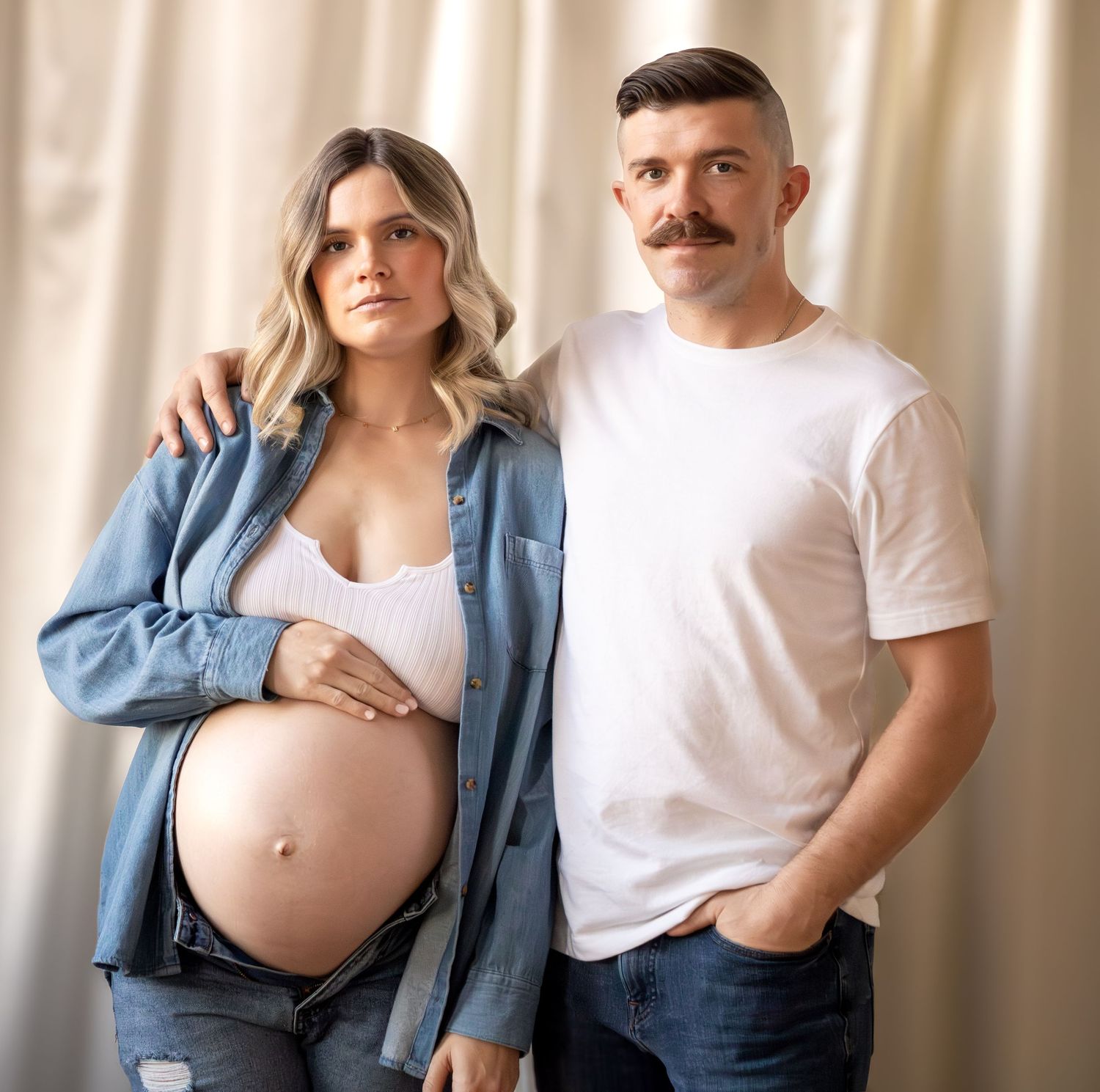 Expectant couple in casual attire poses together against light curtain background during maternity photoshoot.