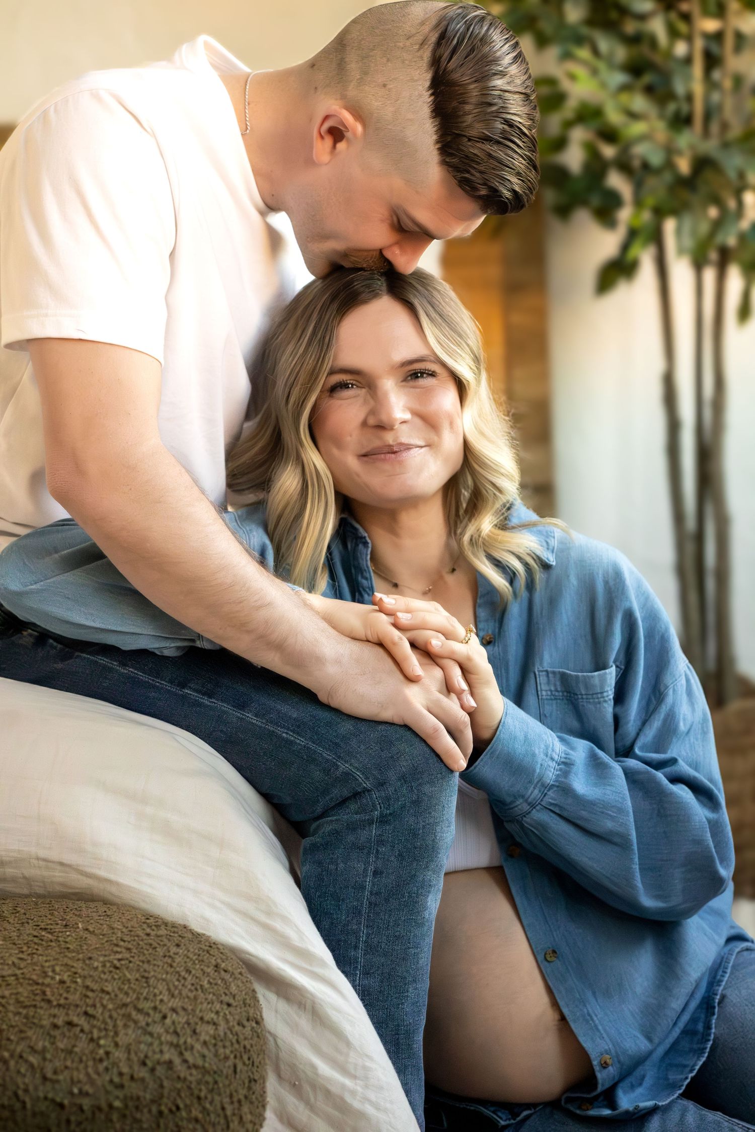 A tender moment between an expecting couple as they sit together indoors near a houseplant.