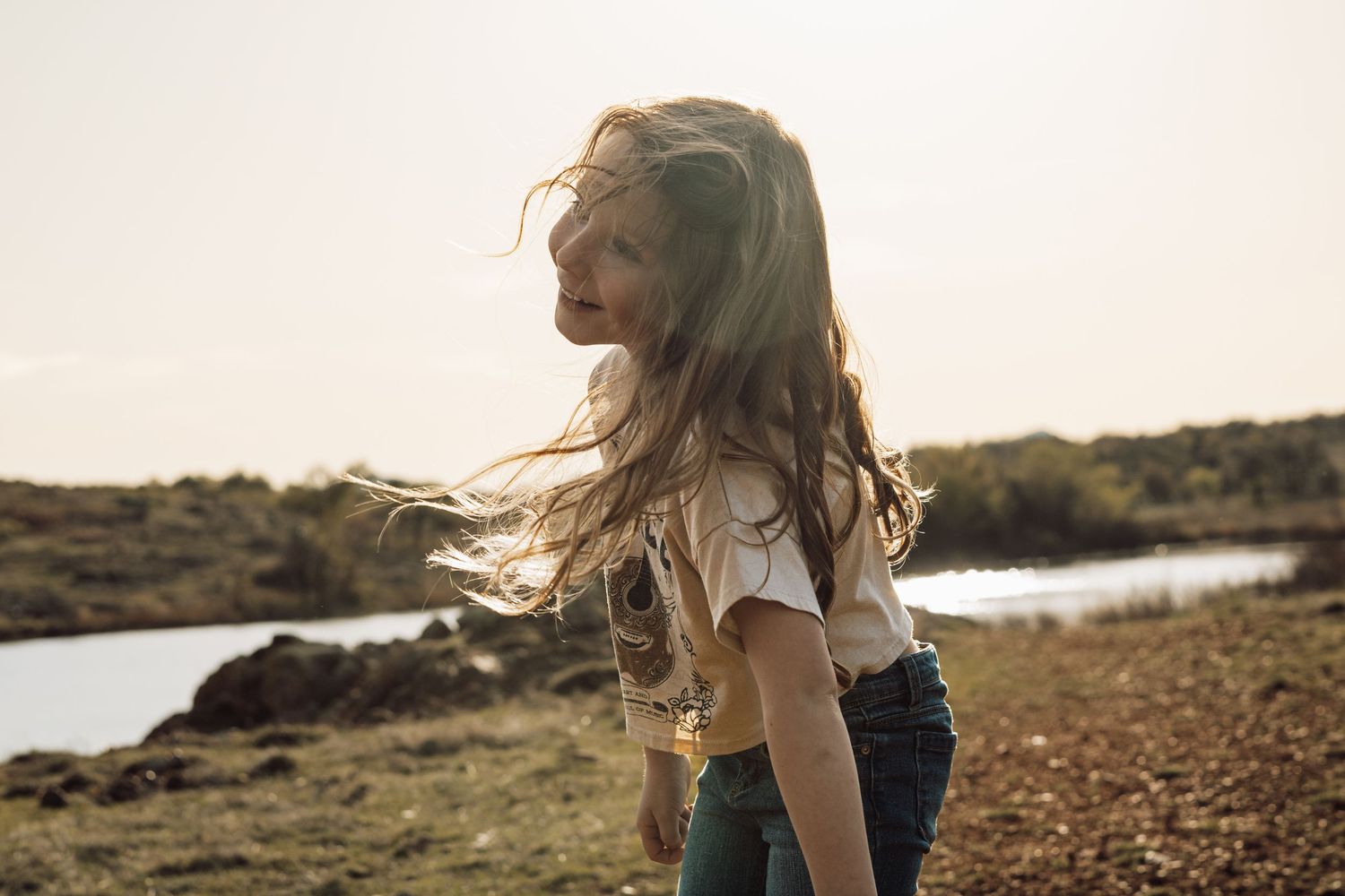 Child with flowing hair walks along lake path at golden hour.