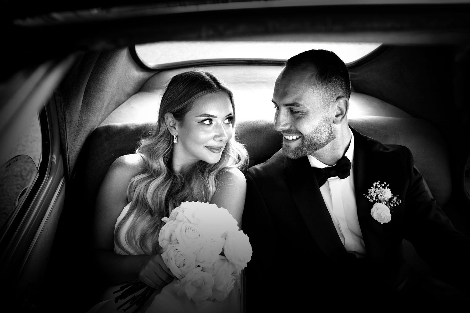 Black and white photo of a bride and groom smiling together in the back seat of a car.