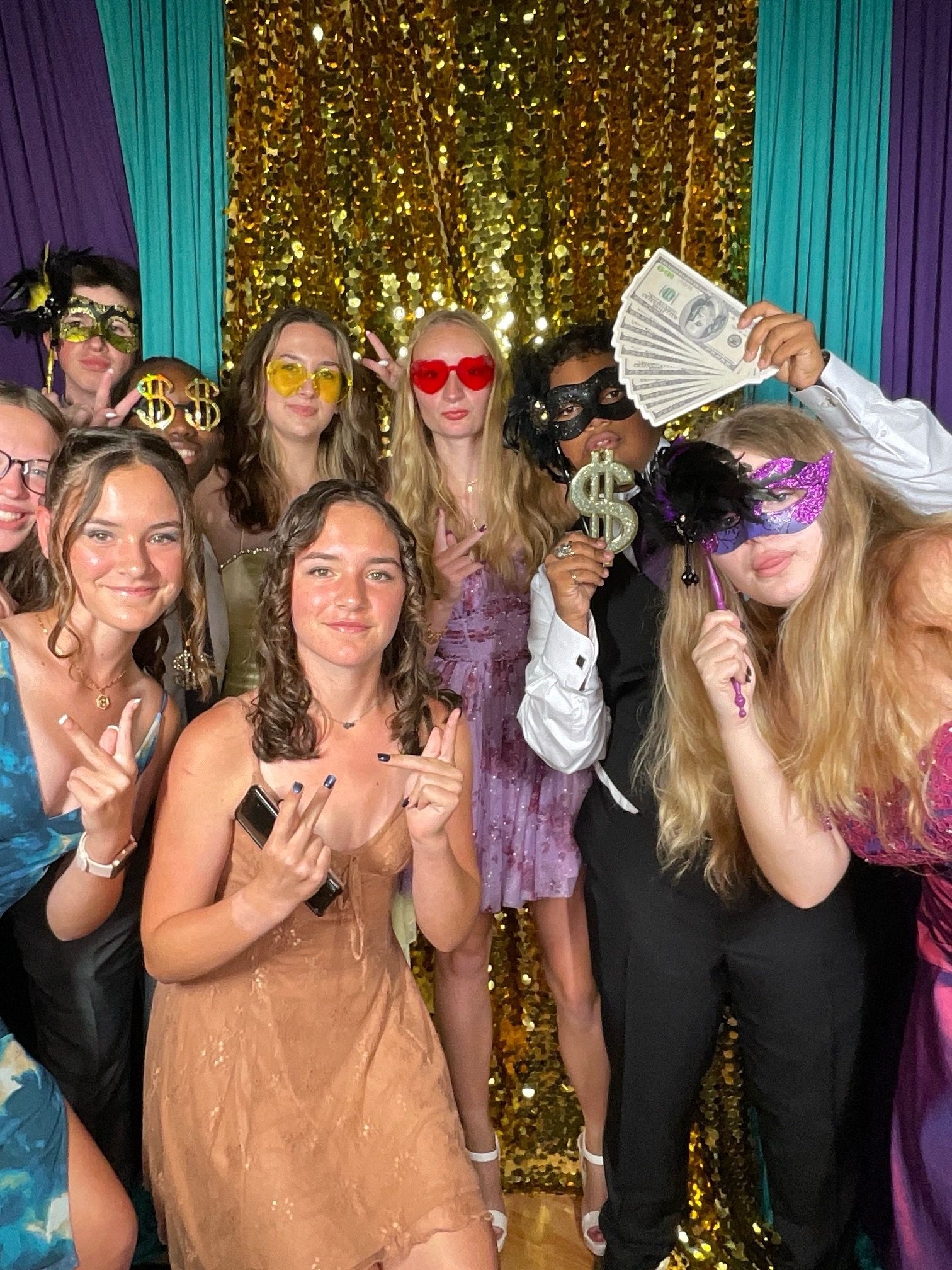 Group of party guests holding up cash and wearing festive accessories against a gold sparkly backdrop.