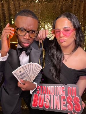 Two people pose with money and sunglasses against a gold sparkly backdrop with text reading "Standing on Business".