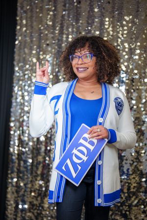 Sorority member in blue and white letterman cardigan holding Zeta sign against sequin backdrop.