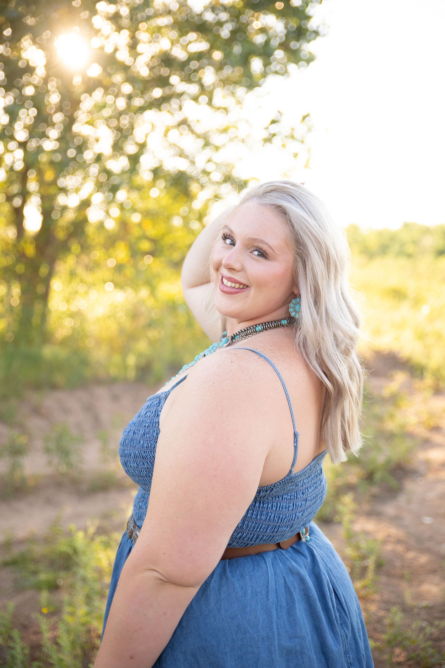 Someone lying in tall grass at sunset wearing a black lace top and jeans while smiling at the camera.
