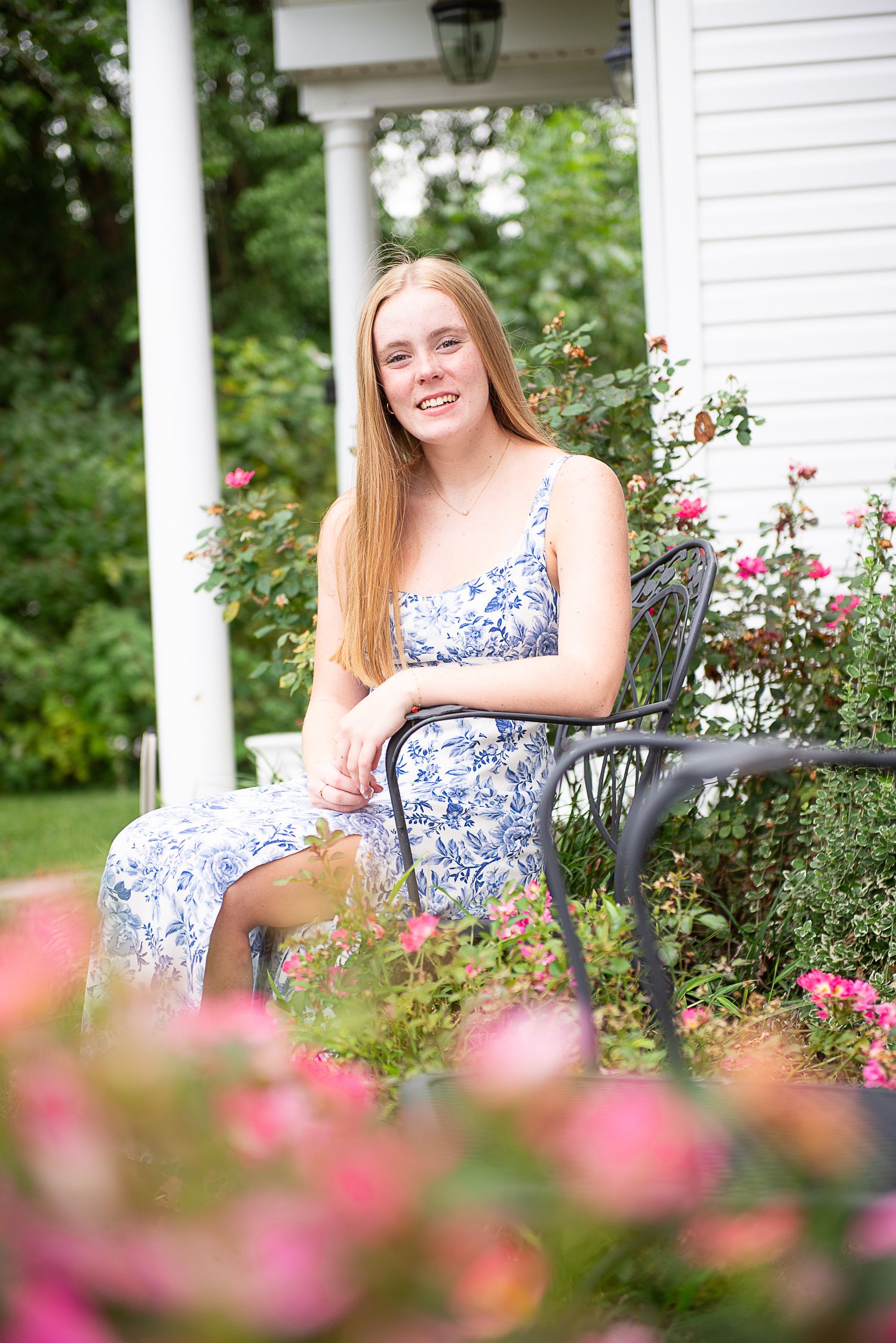 A young woman in a floral dress sits on a porch surrounded by blooming pink roses and white columns.