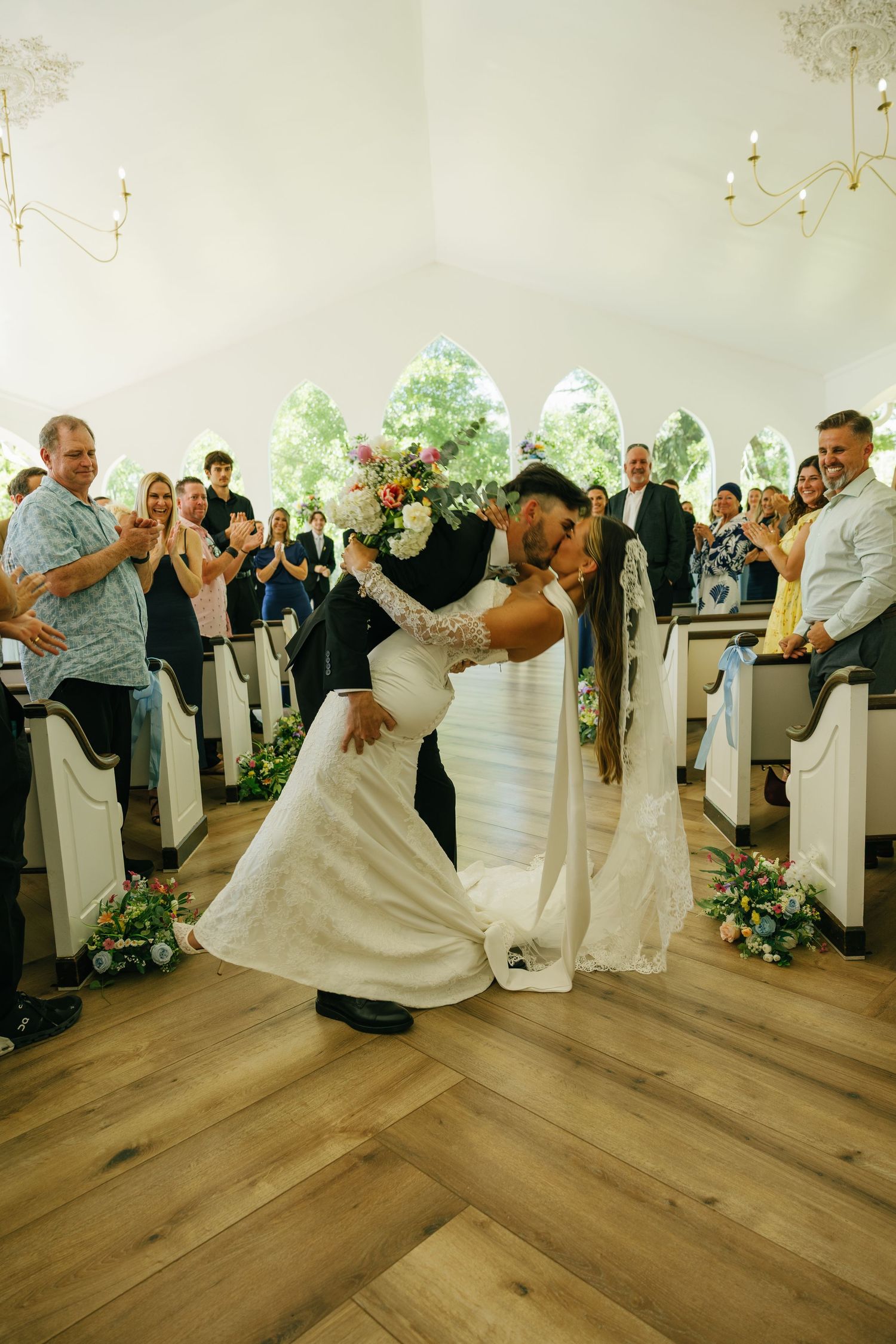 A couple exchanges vows under a draped floral arch at an outdoor wedding ceremony with seated guests watching.