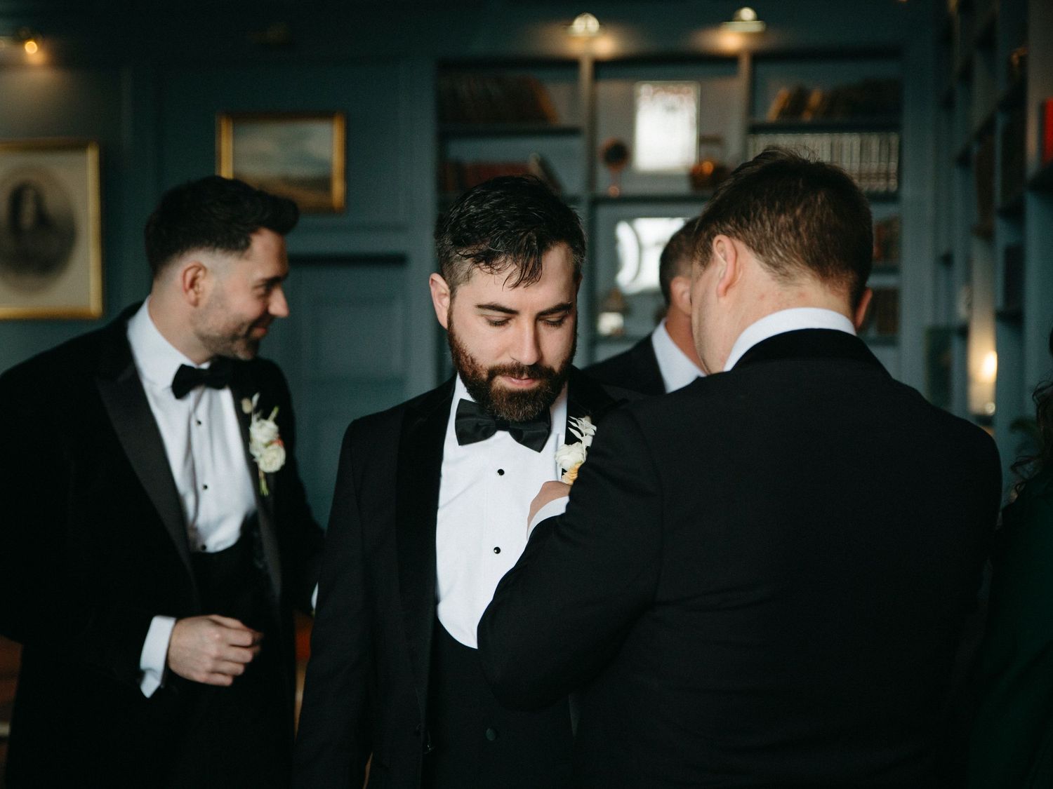 Groomsmen in black tuxedos and bow ties getting ready for a formal event in dim lighting.