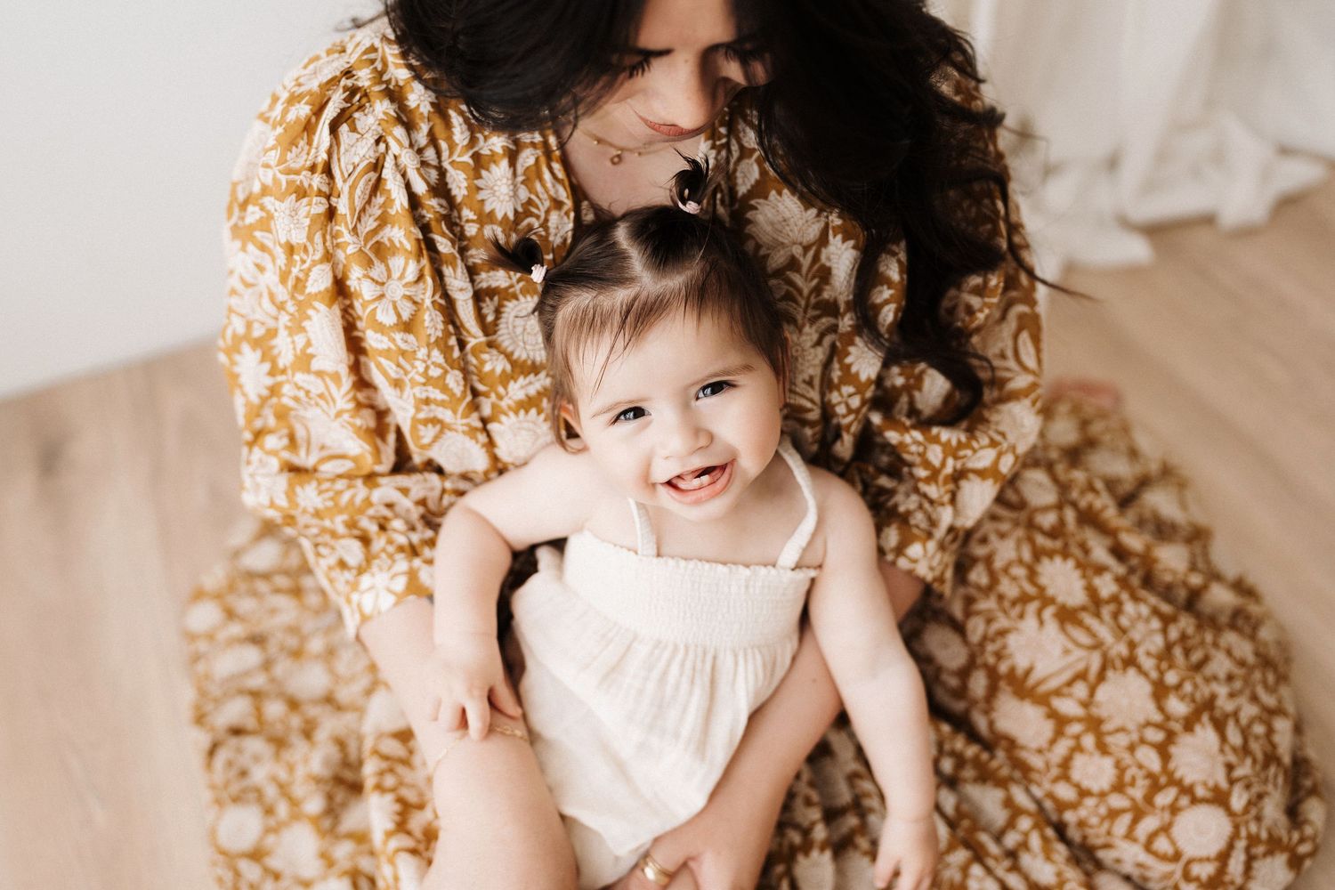 Mother in floral dress embracing smiling toddler in white dress against neutral background.