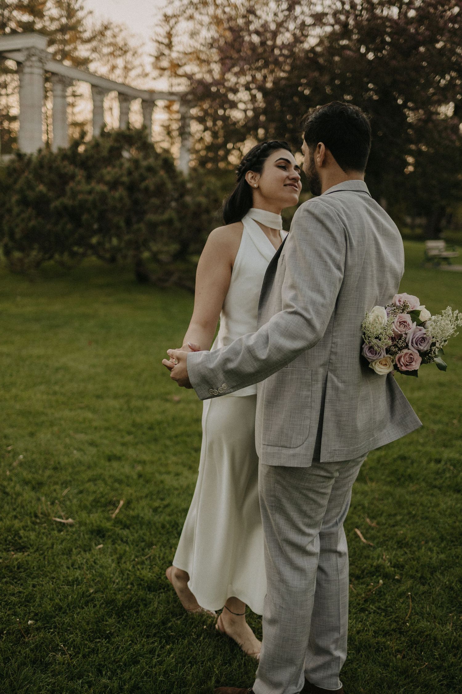 COUPLE AT THE PARK, WITH GREEK MONUMENTS, DANCING. SHE IS HOLDING FLOWERS AND THEY ARE LOOKING AT EACH OTHER,  A SUNSET ON THE BACKGROUND