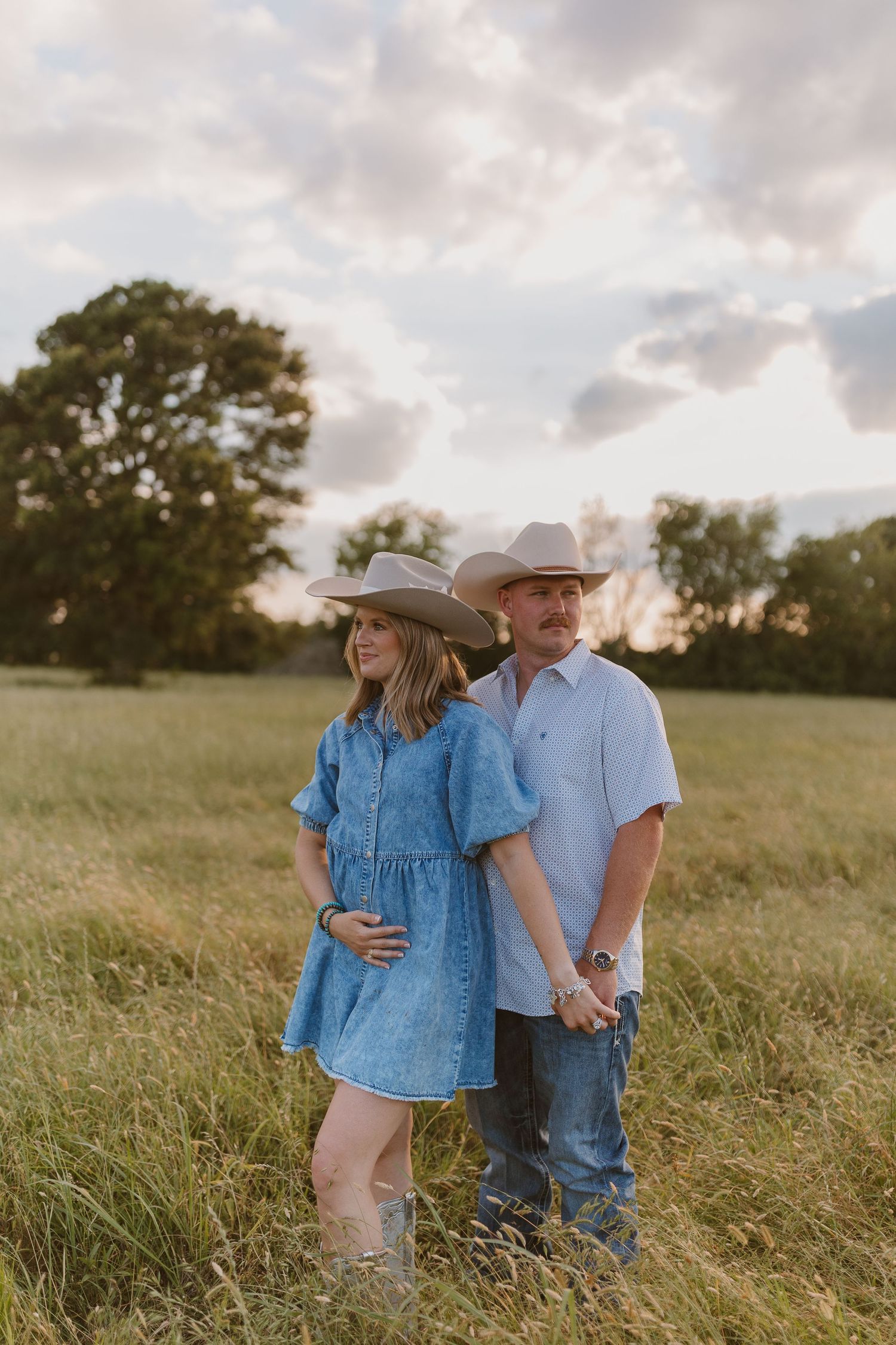 A couple in western attire stands together in a sunlit meadow at golden hour wearing matching blue outfits.