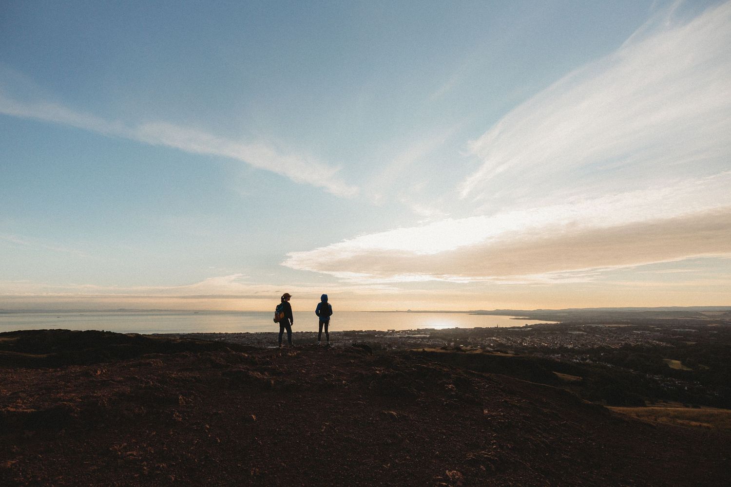 A hiker with two border collies stands on a rocky mountain summit overlooking a distant landscape at sunrise.