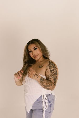 Fashion model in white corset top and light denim poses against neutral background showing sleeve tattoos.