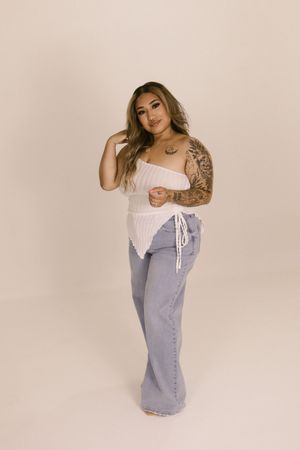 Plus size model poses in white corset and light denim against beige backdrop.