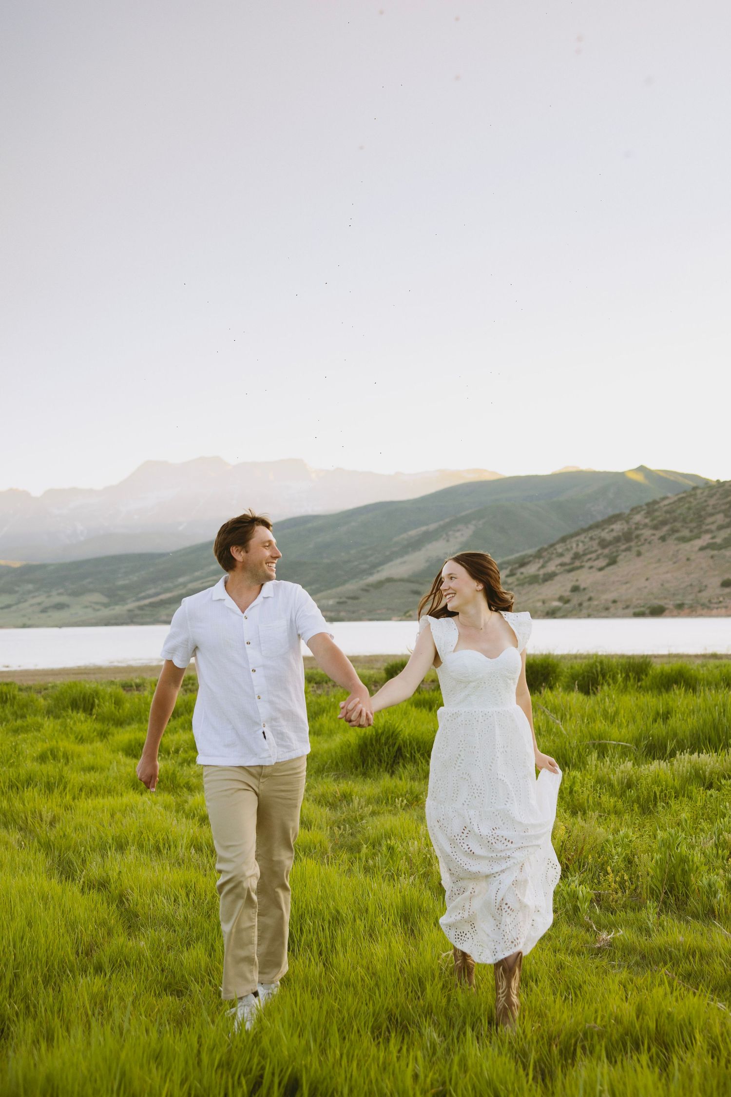 A couple dances in a scenic meadow at sunset with majestic mountains and golden sunlight in the background.