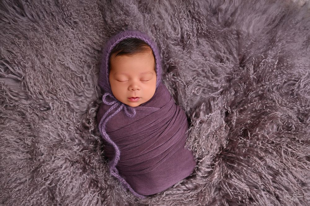 Sleeping newborn wrapped in purple swaddle on textured fur blanket.