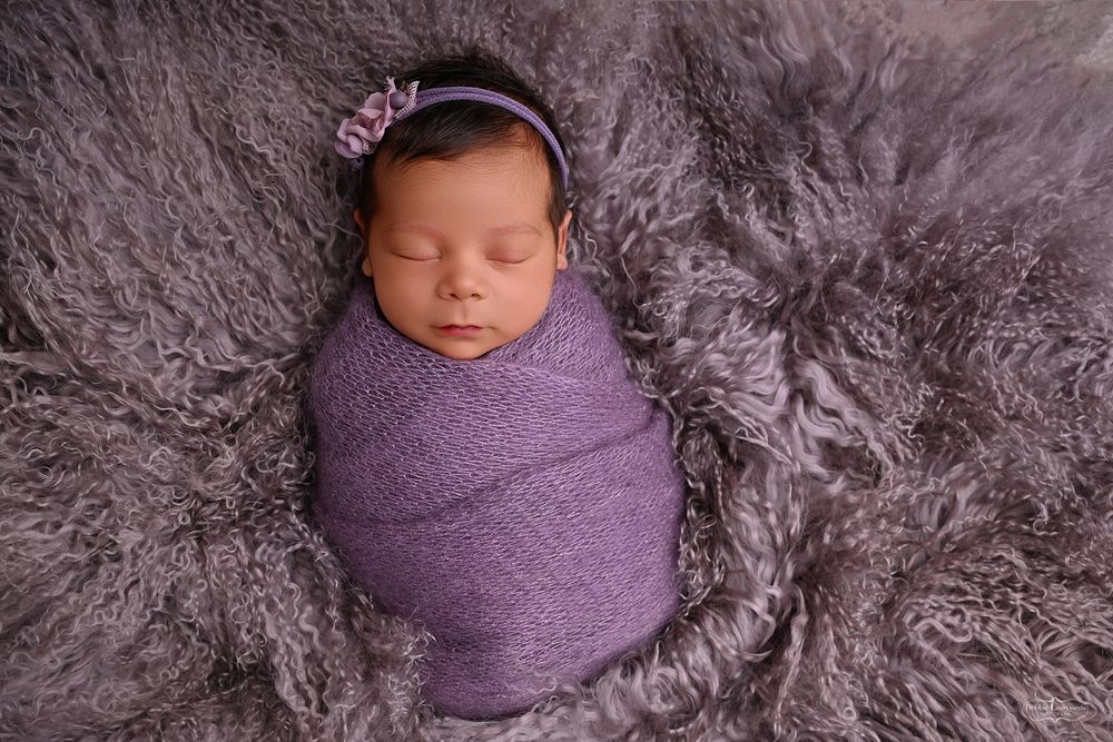 Peaceful newborn wrapped in purple fabric resting on soft fur backdrop.