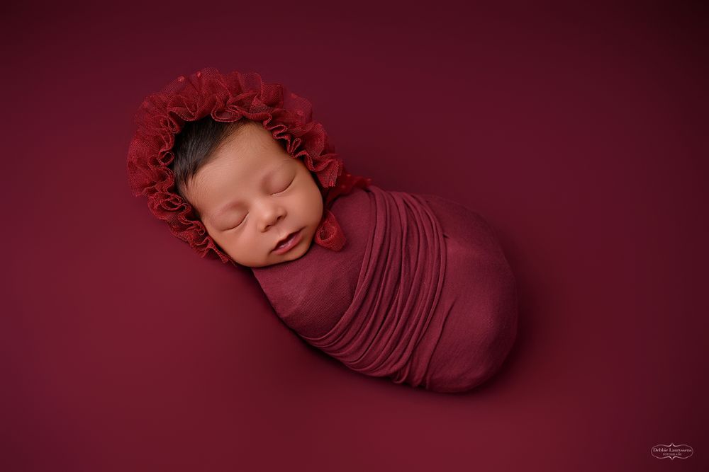 Sleeping newborn with red bonnet wrapped in burgundy fabric against burgundy background.
