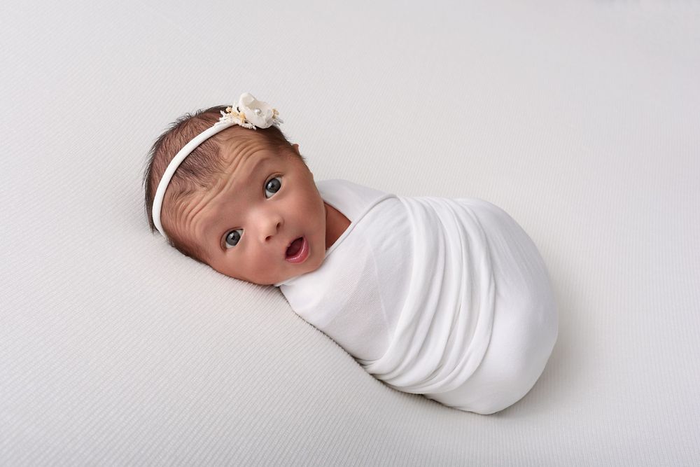 Newborn baby with a lot of expression wrapped in white swaddle with delicate headband against light background.