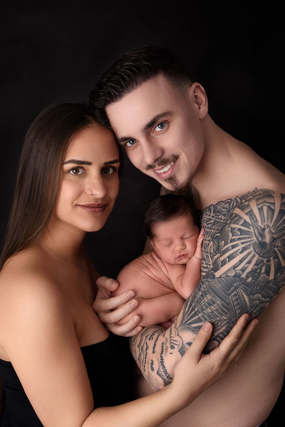 Intimate couple with their newborn photography series against dark background featuring artistic tattoo details.
