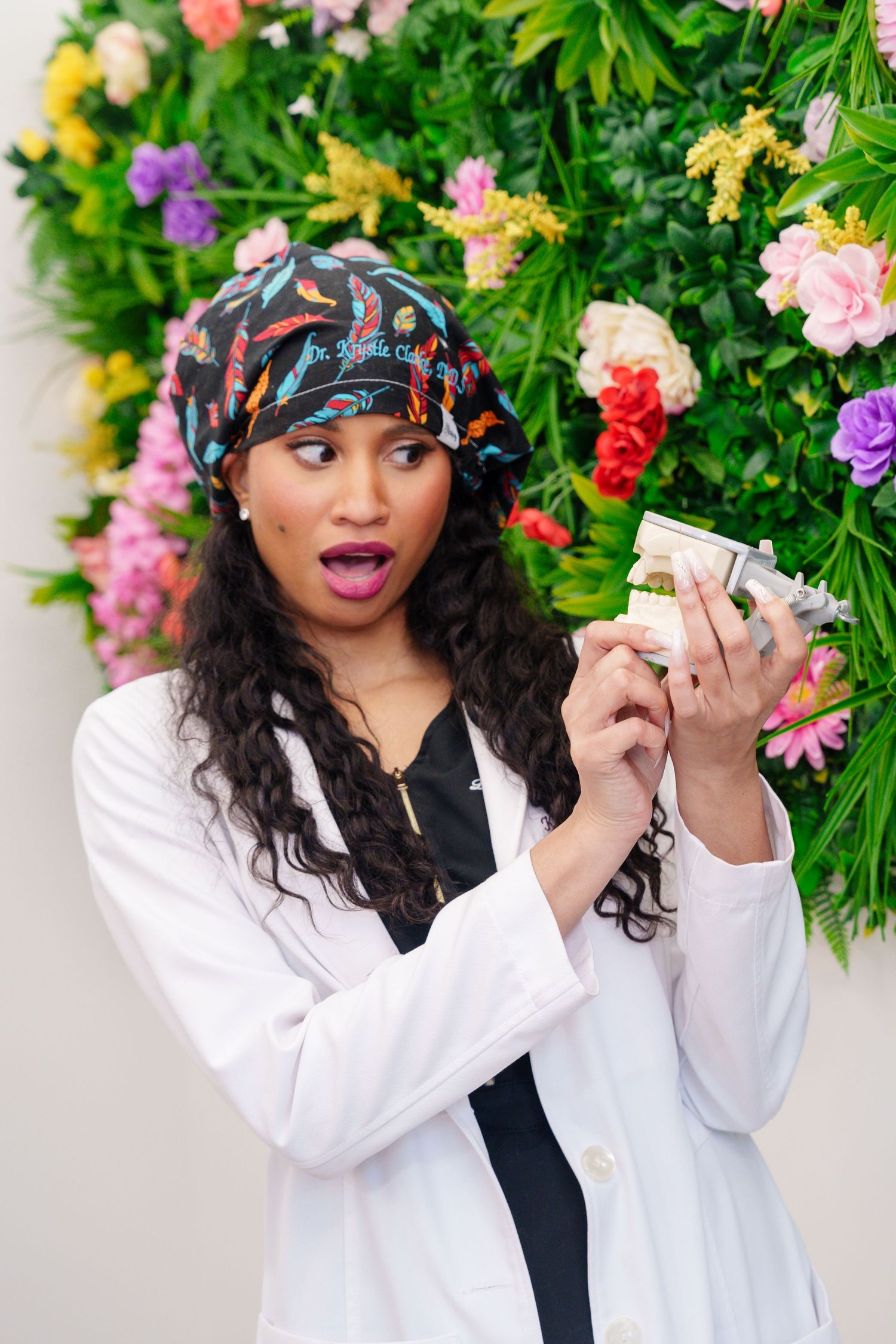 Dr. Clark in a white coat and patterned headscarf shows an excited expression while holding dental sculpture against a floral backdrop in DC